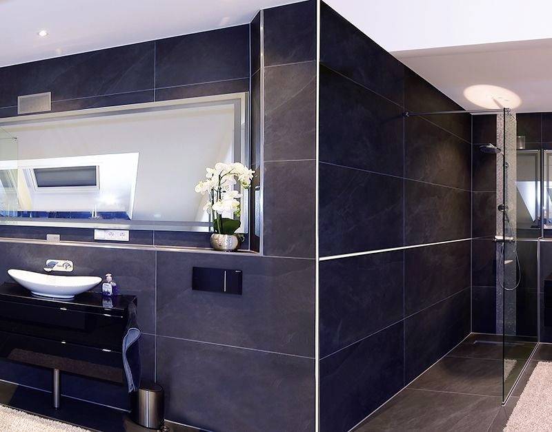 Modernes Badezimmer in dunklen Fliesen, Waschbecken und Spiegel links, Dusche rechts, helle Beleuchtung. Elegante Dekoration mit Orchideen.
