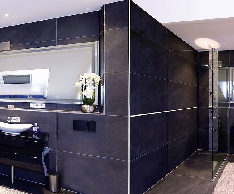 Modernes Badezimmer mit dunklen Fliesen. Links ist ein Waschbecken mit Spiegel, rechts eine ebenerdige Dusche. Orchideen schmücken die Ablage.