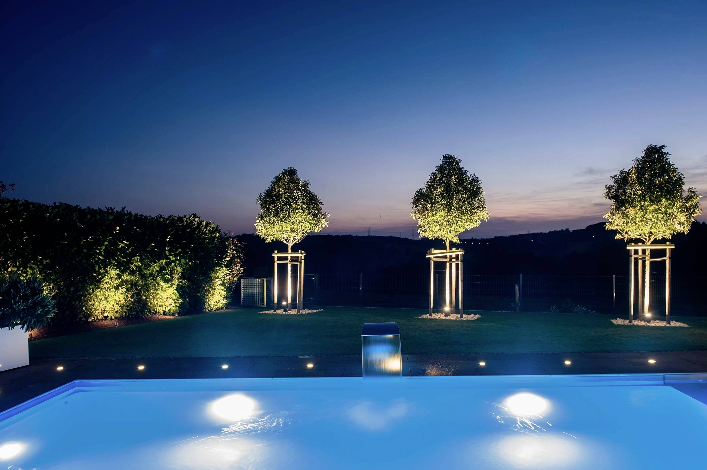 Ein beleuchteter Garten mit drei Bäumen im Hintergrund neben einem Pool, während des Sonnenuntergangs. Die Landschaft ist dunkel und friedlich.