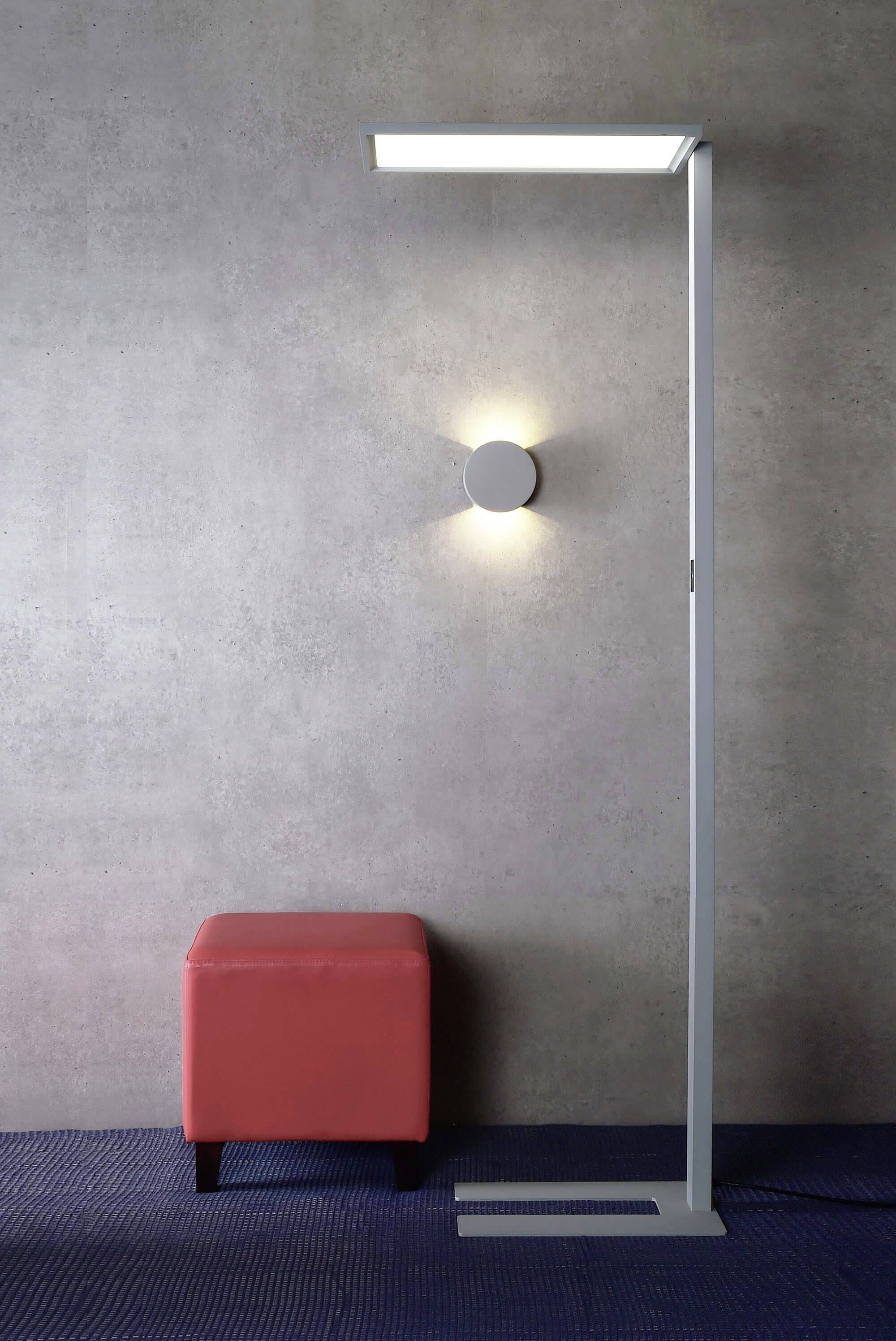 Eine moderne, freistehende Lampe leuchtet eine graue Wand an. Daneben steht ein roter Hocker, der Kontrast zum schlichten Design bietet.