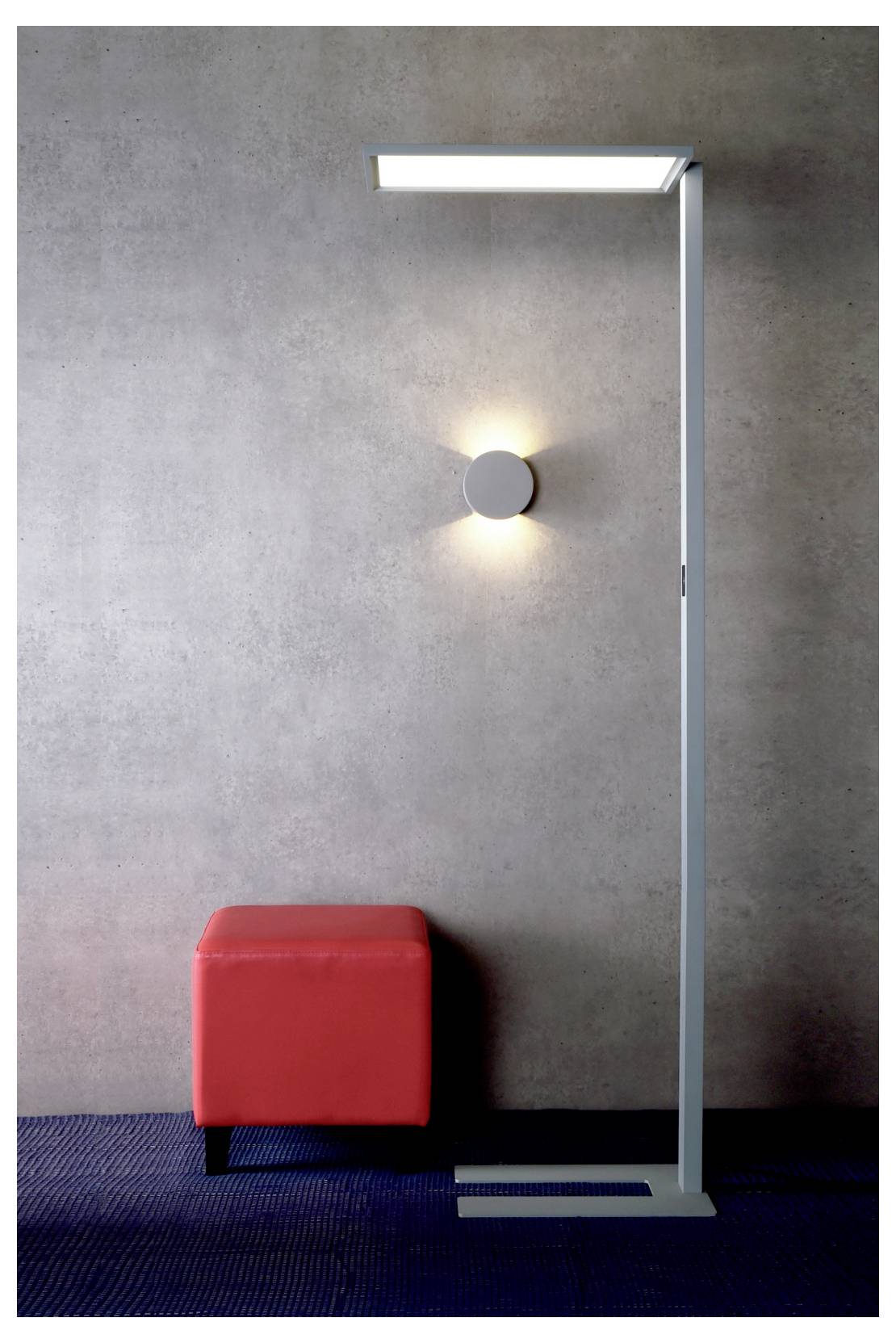 Moderne Inneneinrichtung mit einer hohen Stehlampe neben einem roten Würfelhocker vor einer grauen Wand und einem dunkelblauen Teppich.