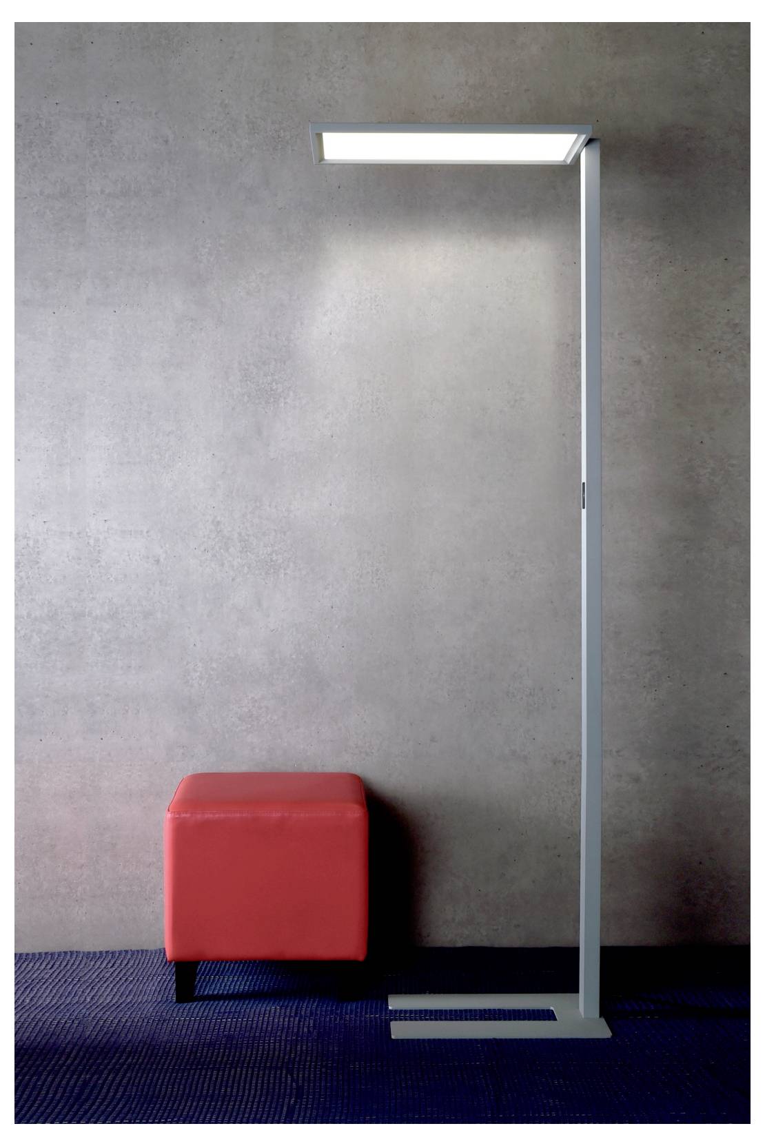 Roter Würfelhocker neben einer hohen, modernen Stehlampe vor einer grauen Wand auf einem dunklen Teppichboden.