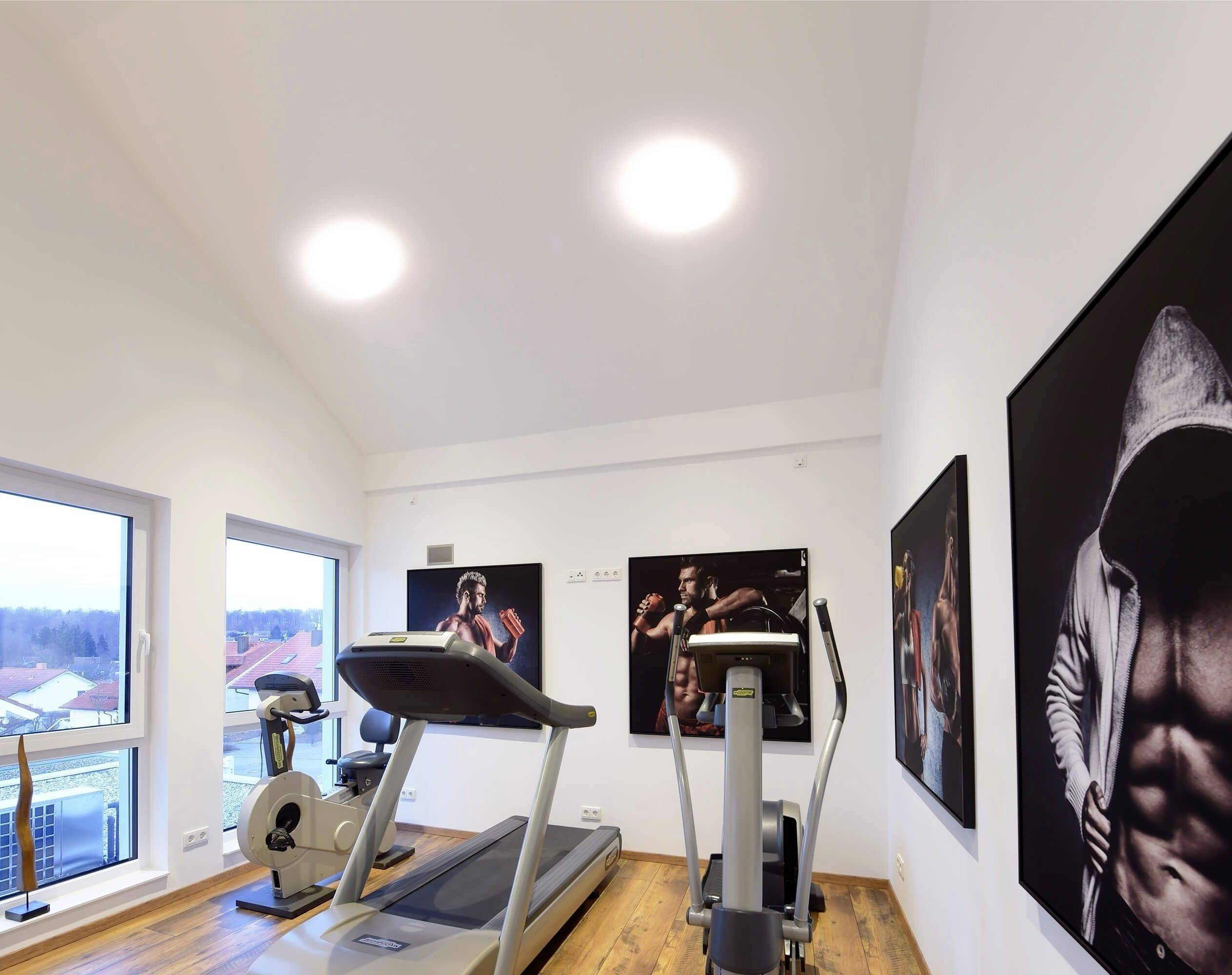 Fitnessraum mit Laufband, Heimtrainer und Boxkunst an den Wänden. Große Fenster bieten Blick auf eine ruhige ländliche Umgebung.
