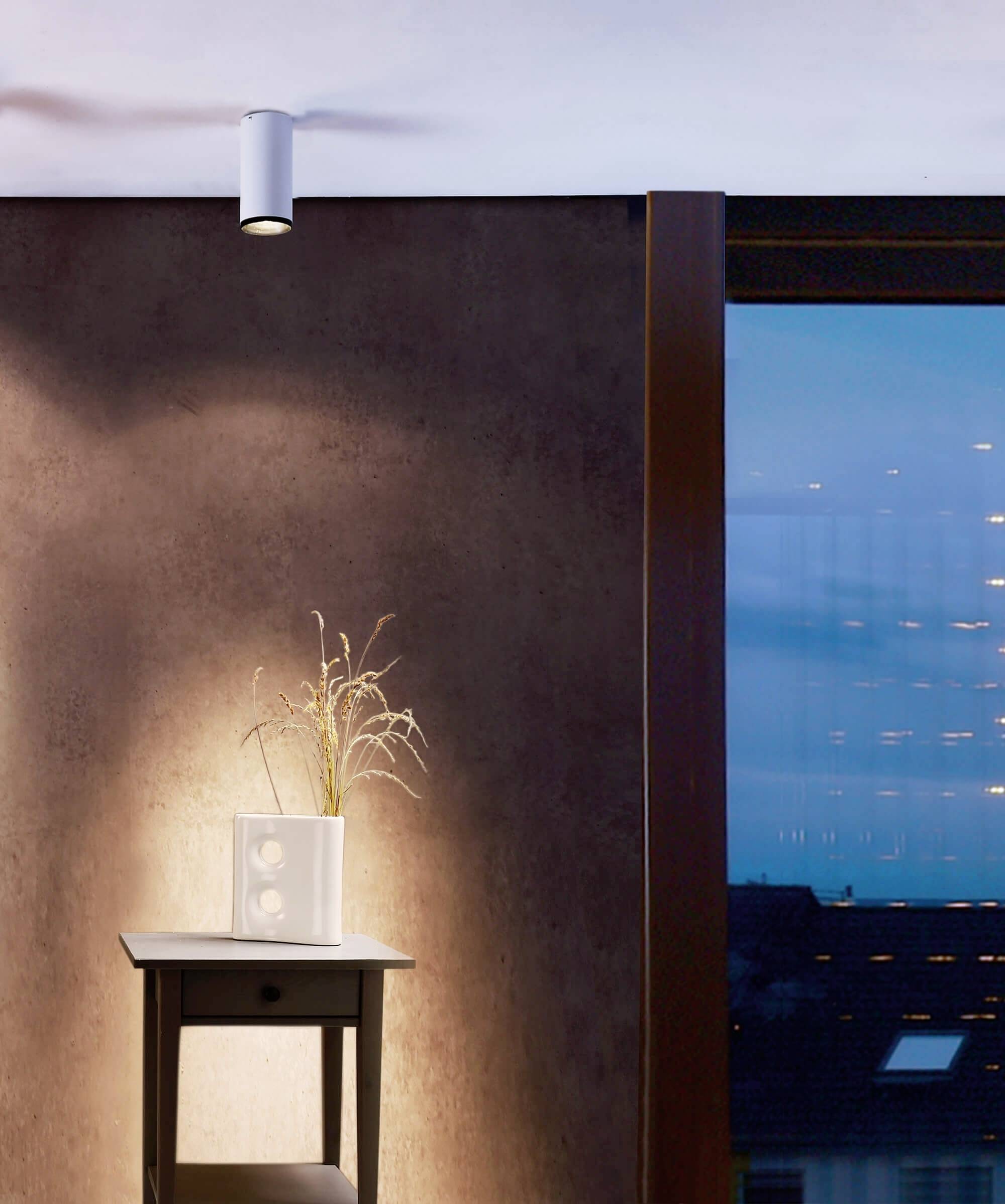 Wandleuchte beleuchtet eine braune Wand mit einem Tisch darunter, auf dem ein Dekoelement steht. Rechts befindet sich ein Fenster mit Abendhimmel.