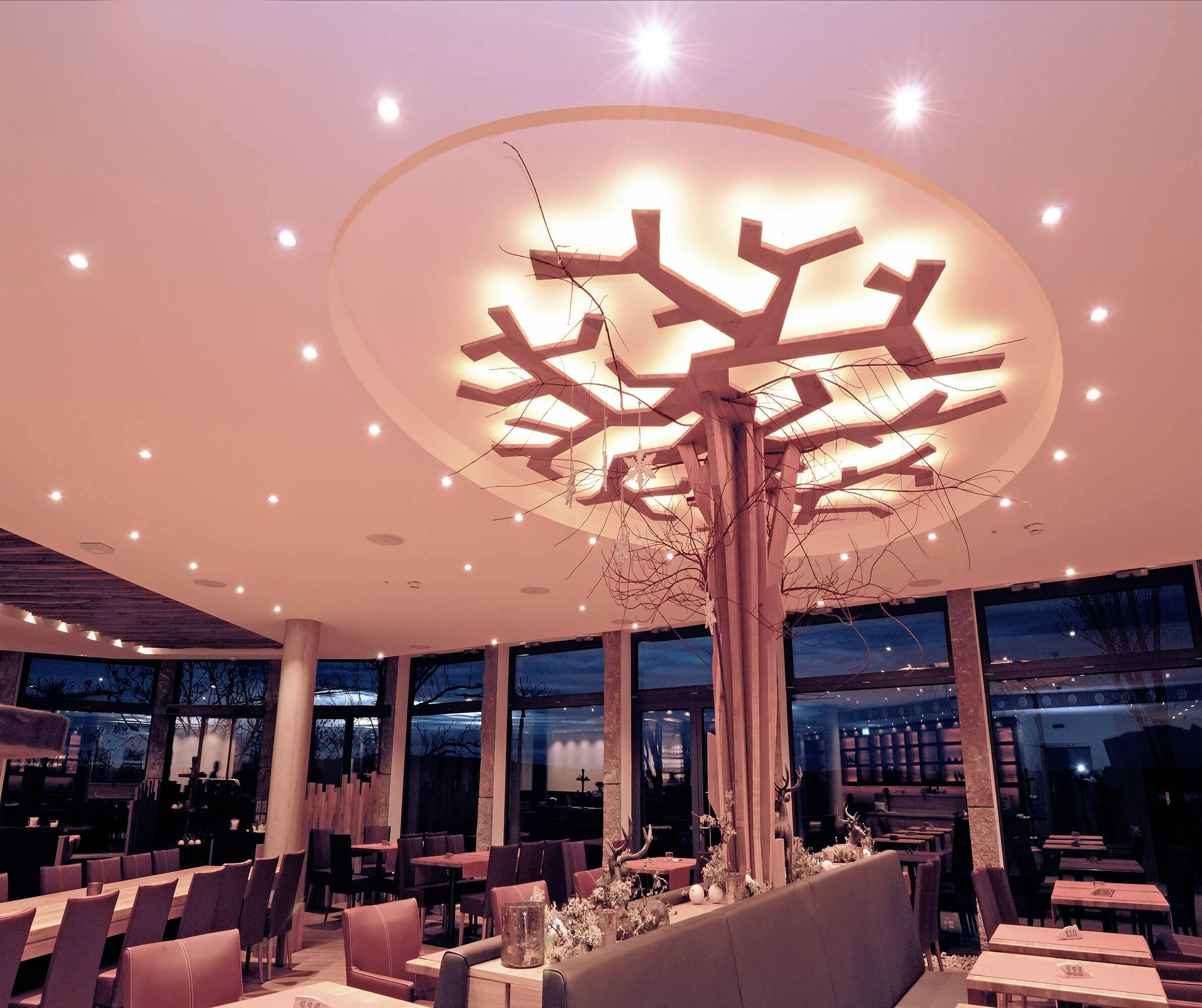 Ein stilvolles Restaurant mit modernem Ambiente. Ein beleuchtetes Baumelement an der Decke dominiert den Raum. Dezentes, warmes Licht.