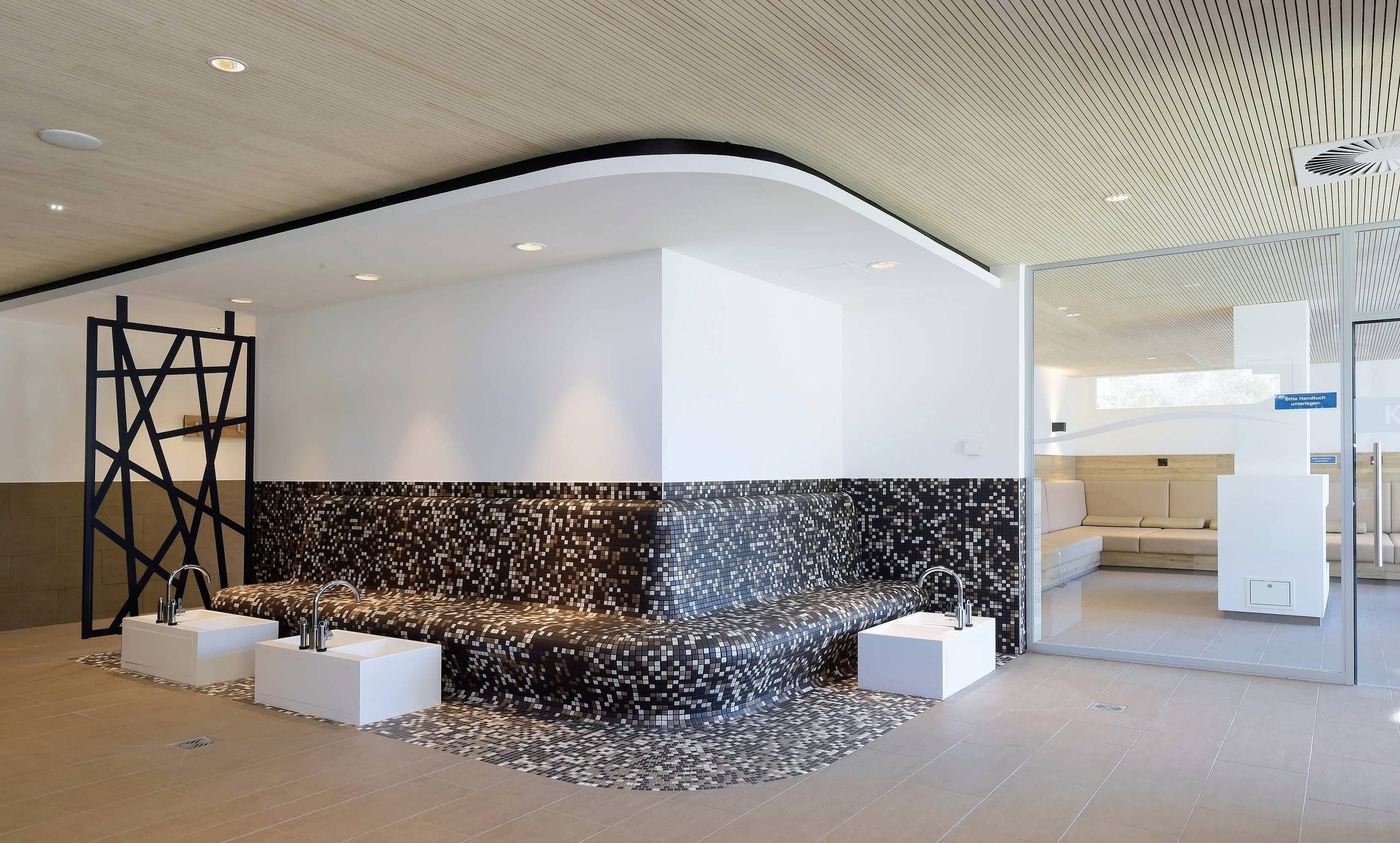 Moderner Entspannungsbereich mit einem dunklen Mosaikbrunnen und weißen Sitzelementen. Im Hintergrund ein Lounge-Bereich.