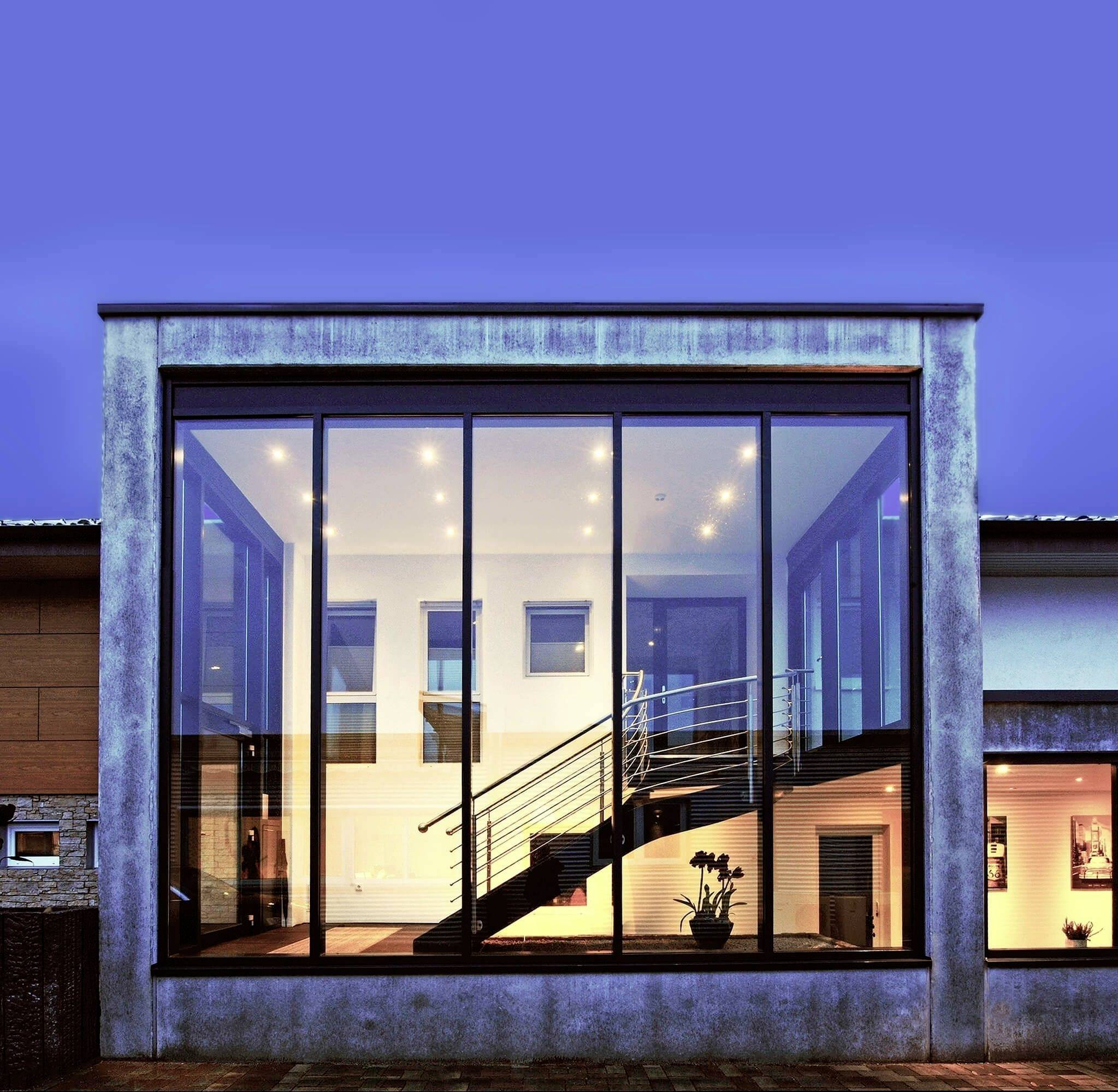 Modernes Gebäude mit großer Glasfront, durch die eine Treppe zu sehen ist. Innen ist warm beleuchtet, kontrastiert mit dunklem Abendhimmel.