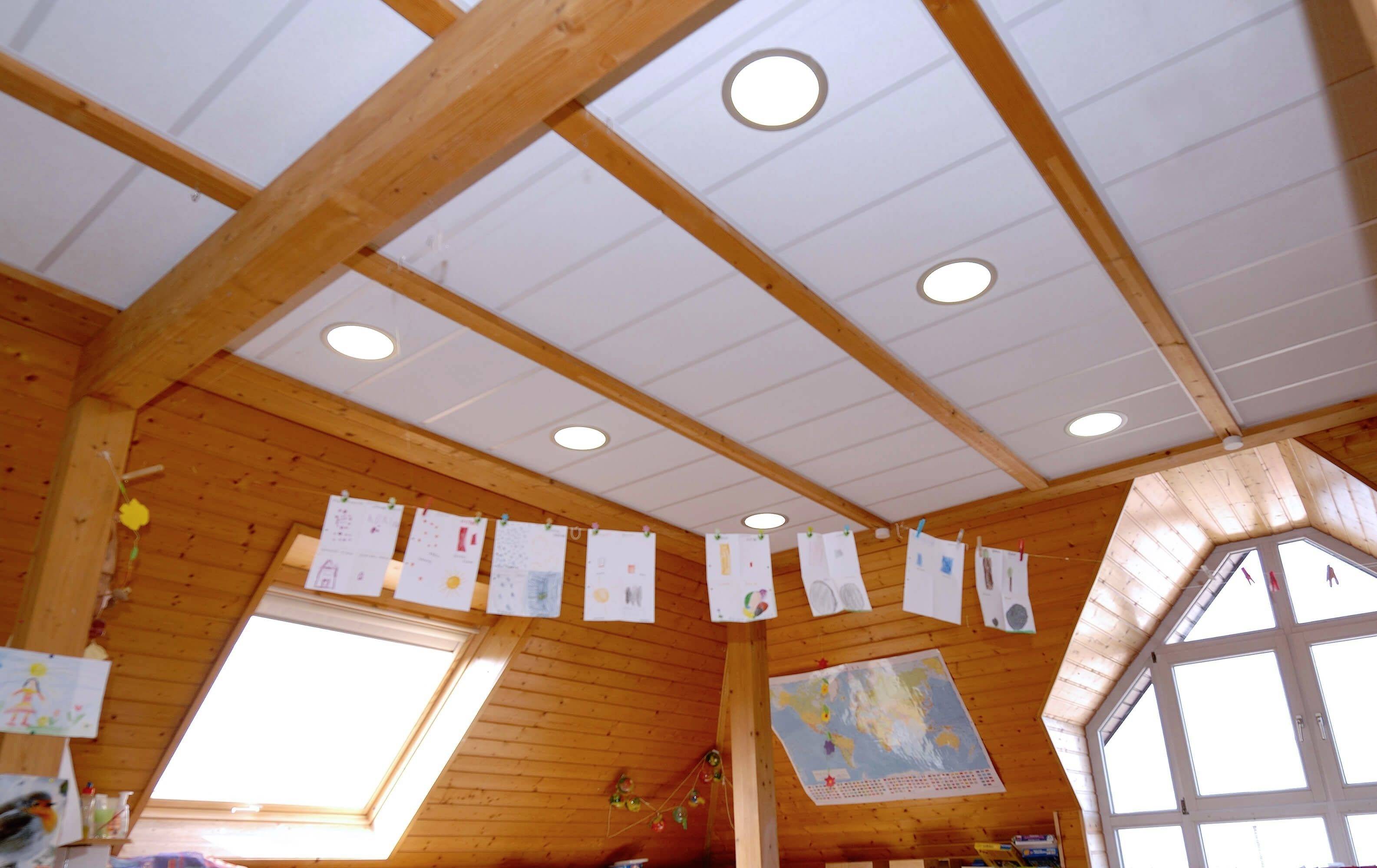 Holzdecke mit mehreren Deckenspots, darunter hängen Kinderzeichnungen an einer Schnur. Rechts ist ein großes Fenster sichtbar.
