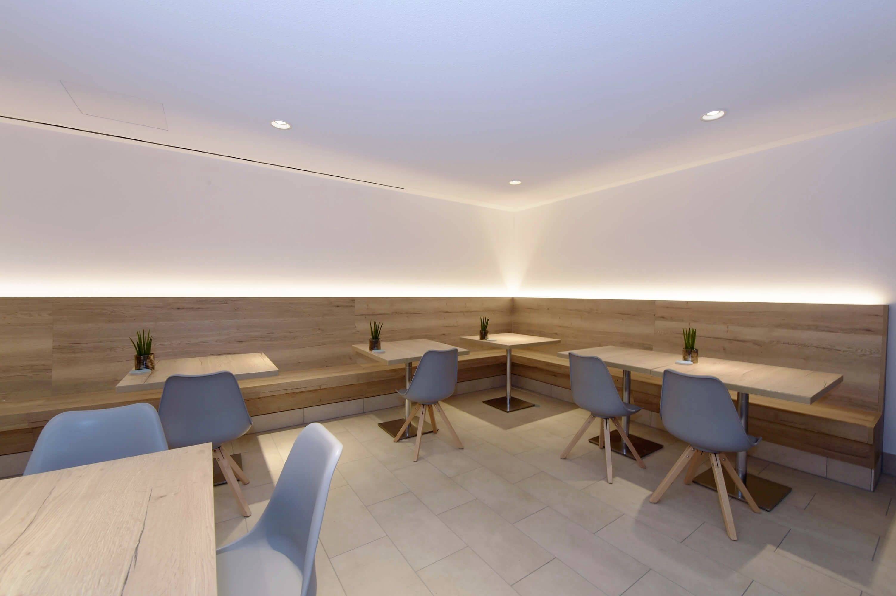 Moderner Café-Raum mit hellen Holz- und weißen Wänden, blau-grauen Stühlen und kleinen Pflanzen in der Ecke der Tische.