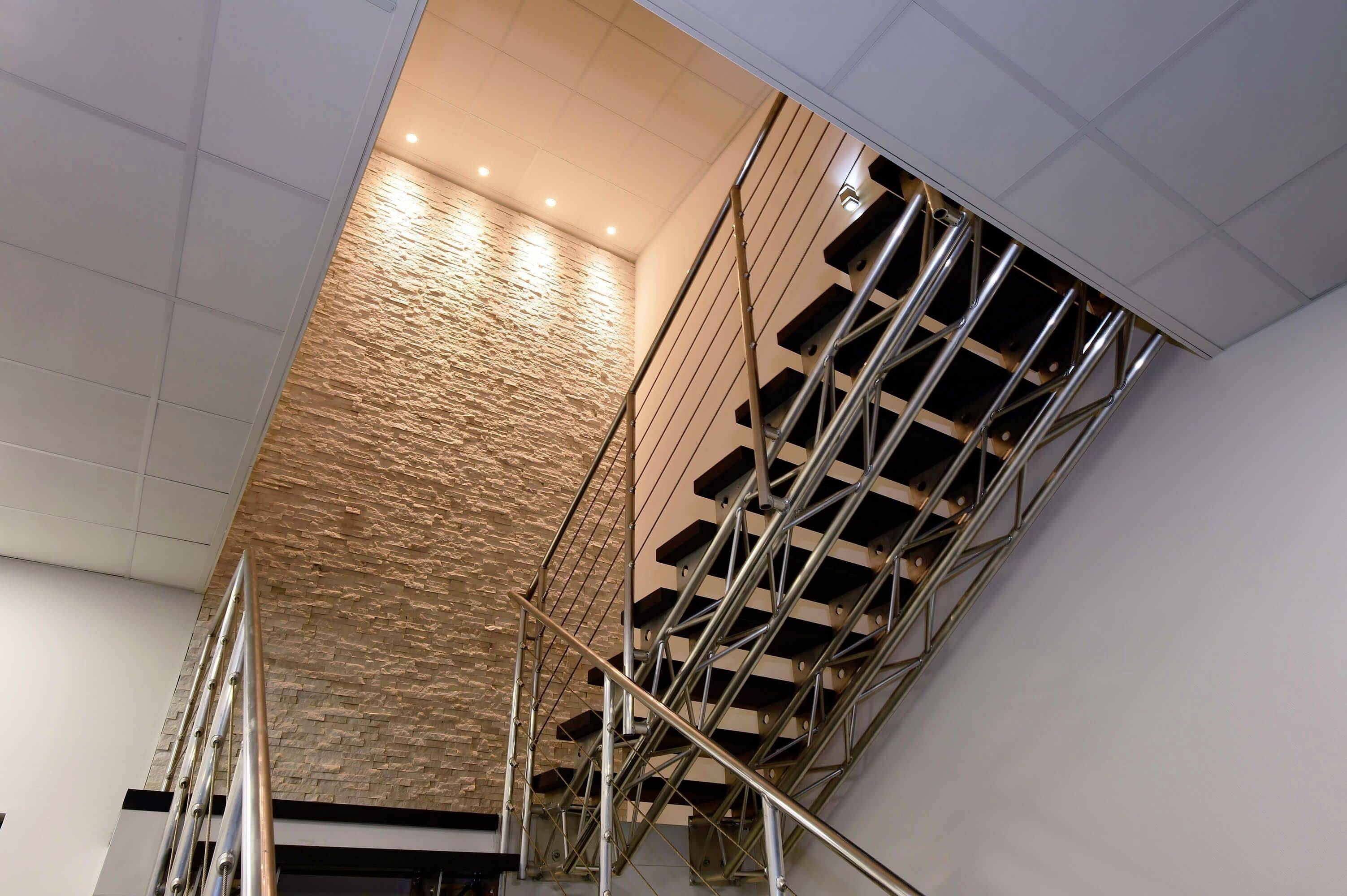 Stahl- und Holztreppe führt zu einer oberen Etage, beleuchtet durch Deckenstrahler. Steinwand bildet den Hintergrund.