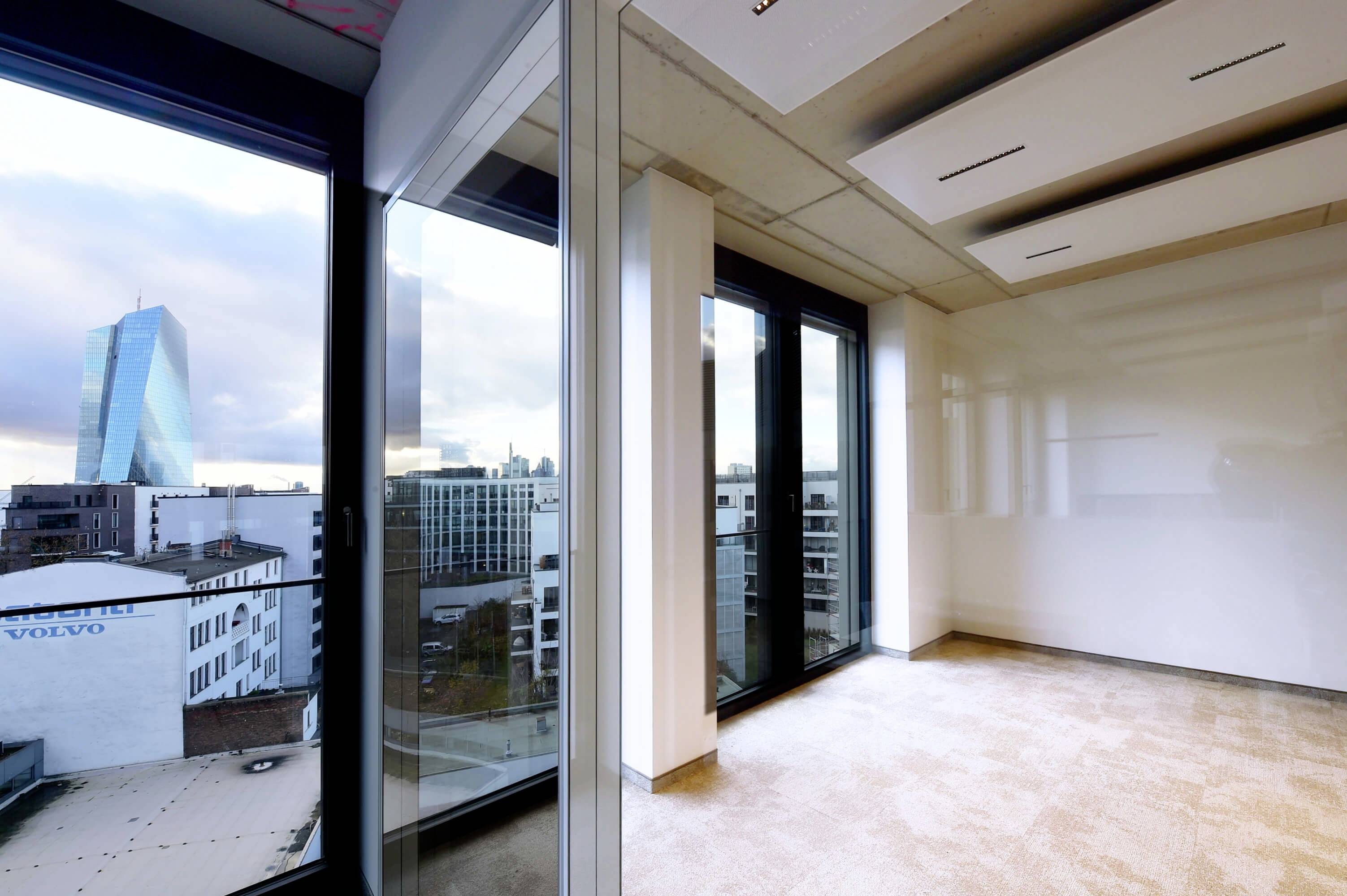 Leeres, modernes Büro mit großen Fenstern, Blick auf die Skyline einer Stadt mit einem markanten Hochhaus im Hintergrund.