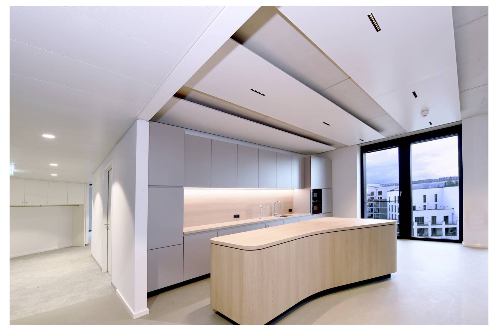 Moderne, minimalistische Küche mit einer geschwungenen Kücheninsel aus hellem Holz, Einbauschränken und einem großen Fenster mit Blick auf Wohnhäuser.