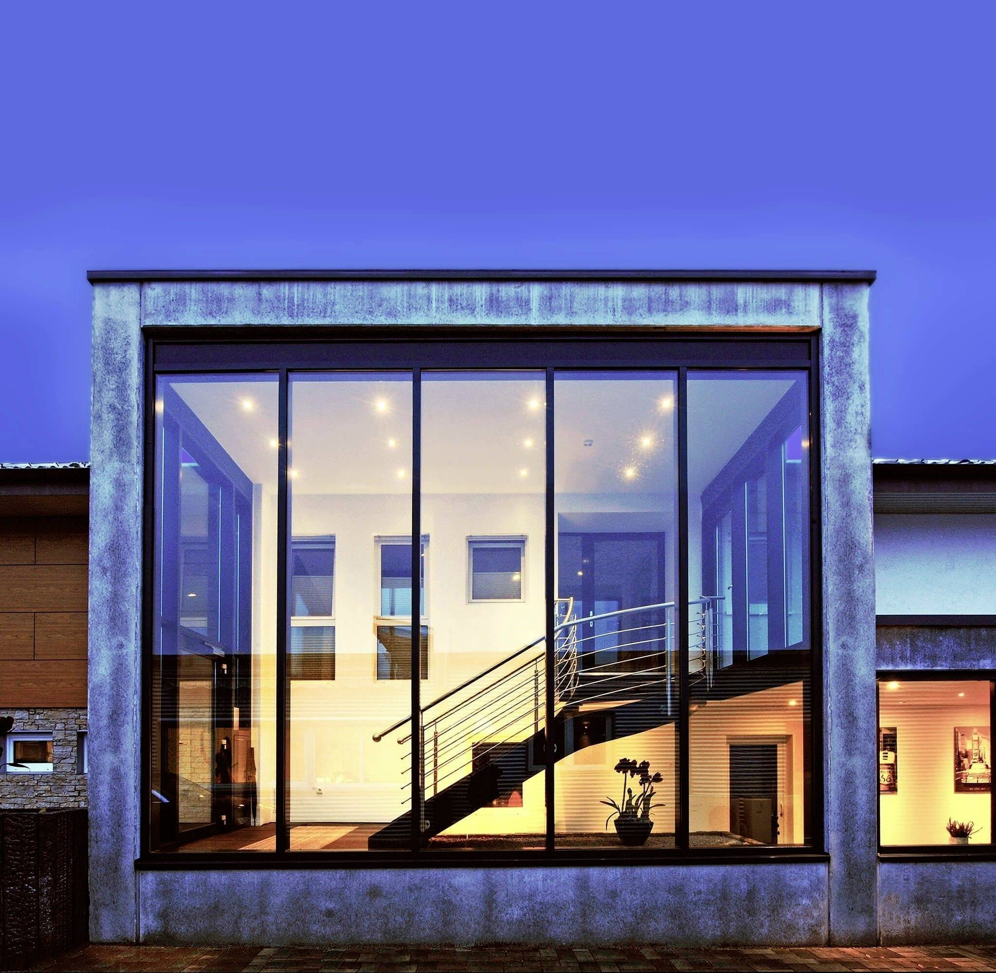 Modernes Gebäude mit breiter Glasfassade. Innen sichtbar, eine Treppe und beleuchtete Räume. Abendszene mit blauem Himmel.