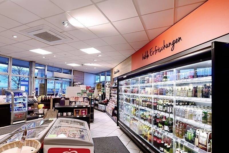 Ein gut beleuchteter Supermarkt mit Regalen voller Getränke und Snacks. Kühlregale rechts, Eingang mit großen Fenstern links im Hintergrund.