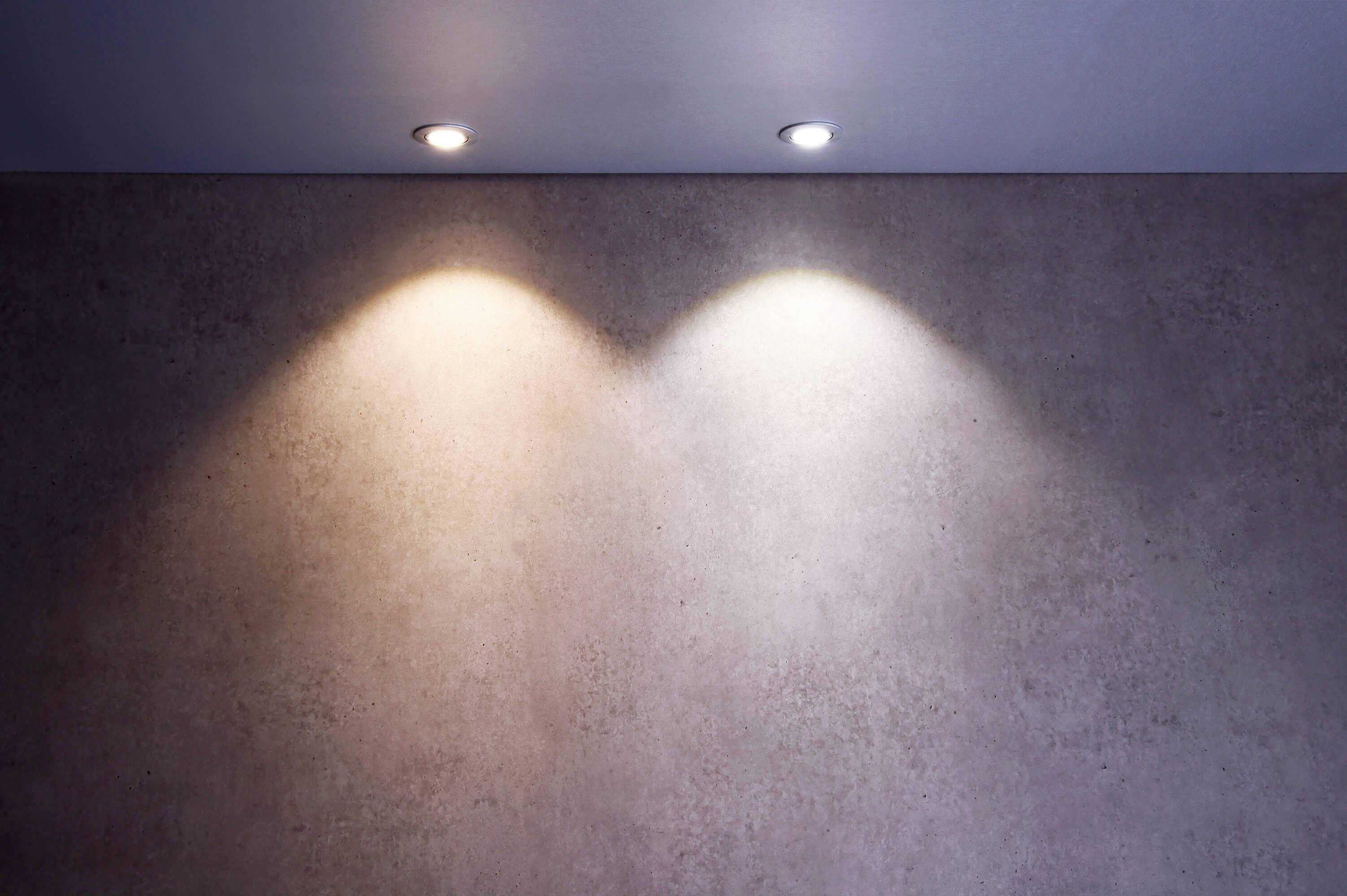 Zwei Deckenleuchten projizieren unterschiedliches Licht auf eine beige Wand, eine warm und gelblich, die andere kühl und weißlich.