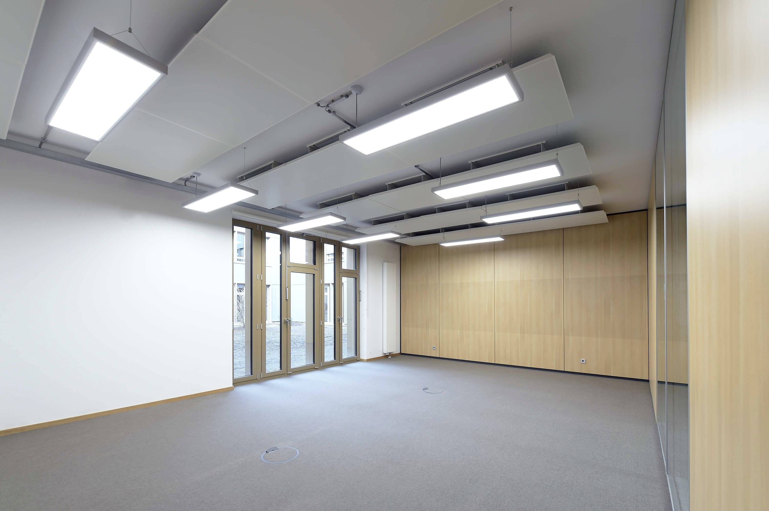 Ein leerer Raum mit grauem Teppichboden, modernen Deckenleuchten und großen Fenstern. Die Wände sind holzverkleidet.