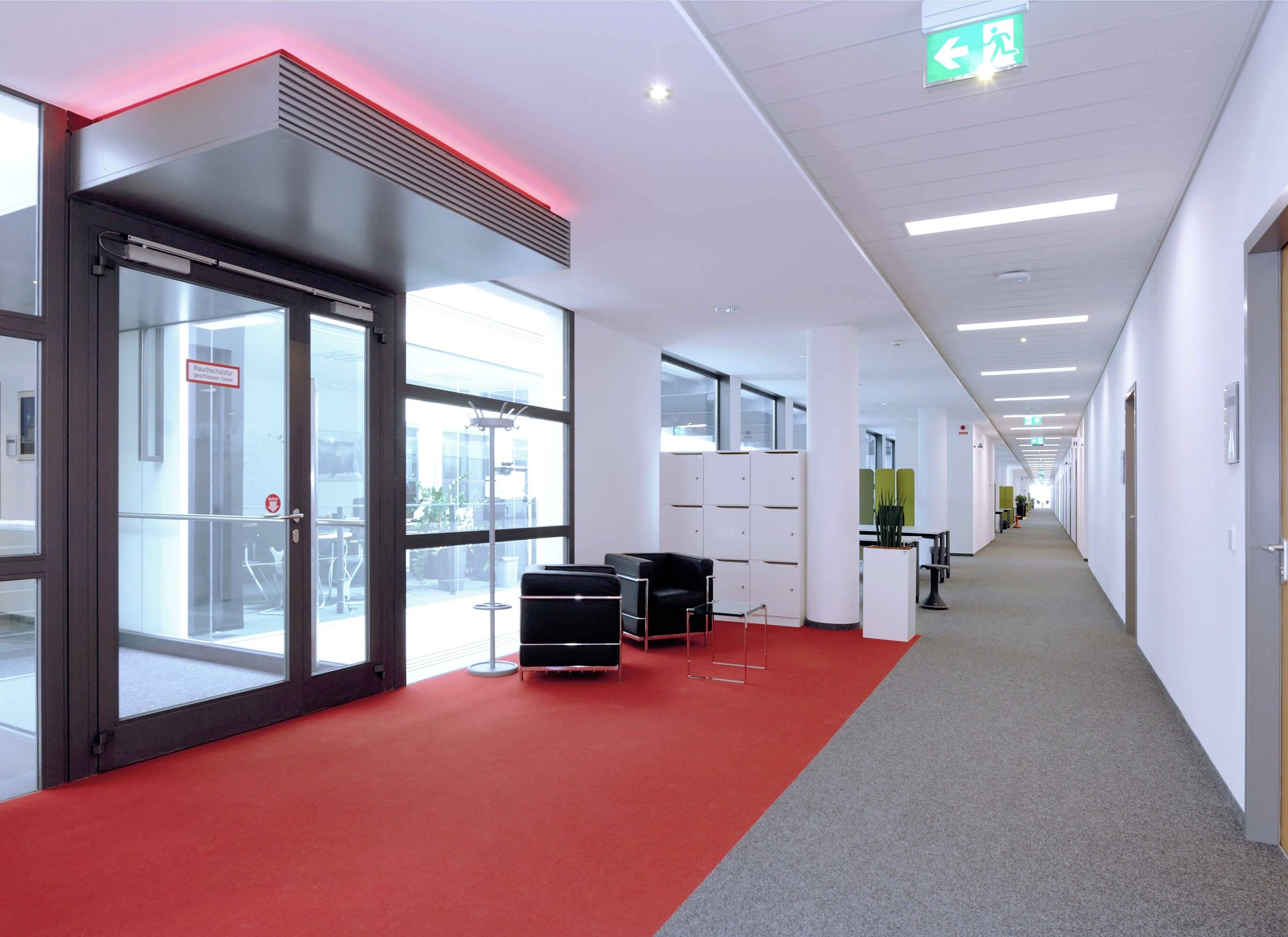 Langer Bürokorridor mit rotem Teppich, Glastüren am Ende und Sitzbereich links. Deutliche Notausgangszeichen an der Decke.