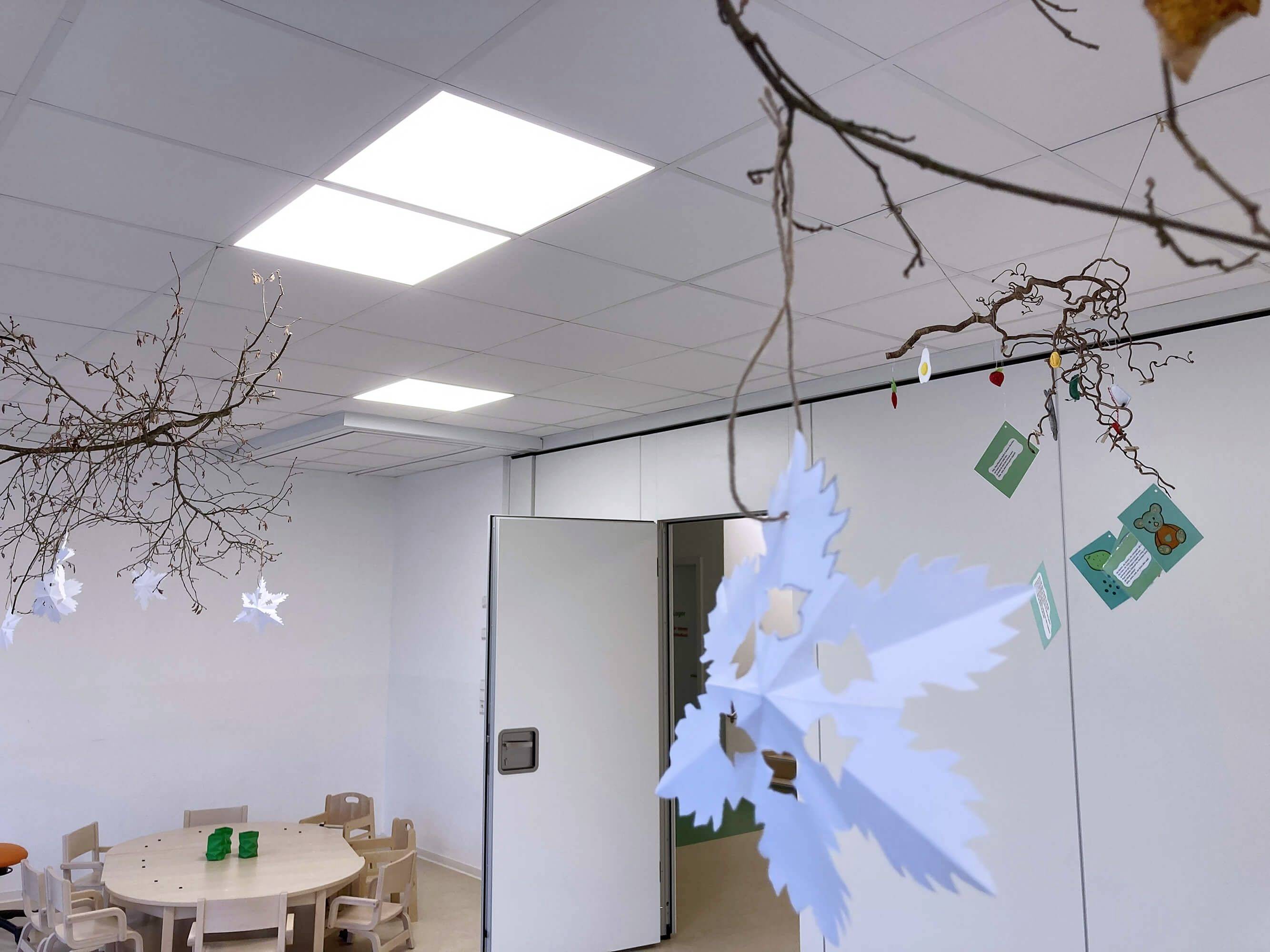 Ein heller Raum mit einem runden Tisch und kleinen Stühlen. An der Decke hängen dekorative Äste mit Papiersternen und Karten.