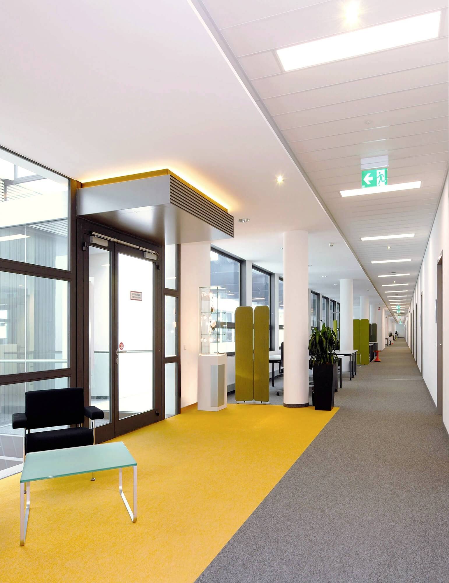 Heller Büroflur mit gelbem Teppich, Glaswänden und einem Ausgang hinten links. Vereinzelt Bürostühle und Pflanzen entlang des Korridors.