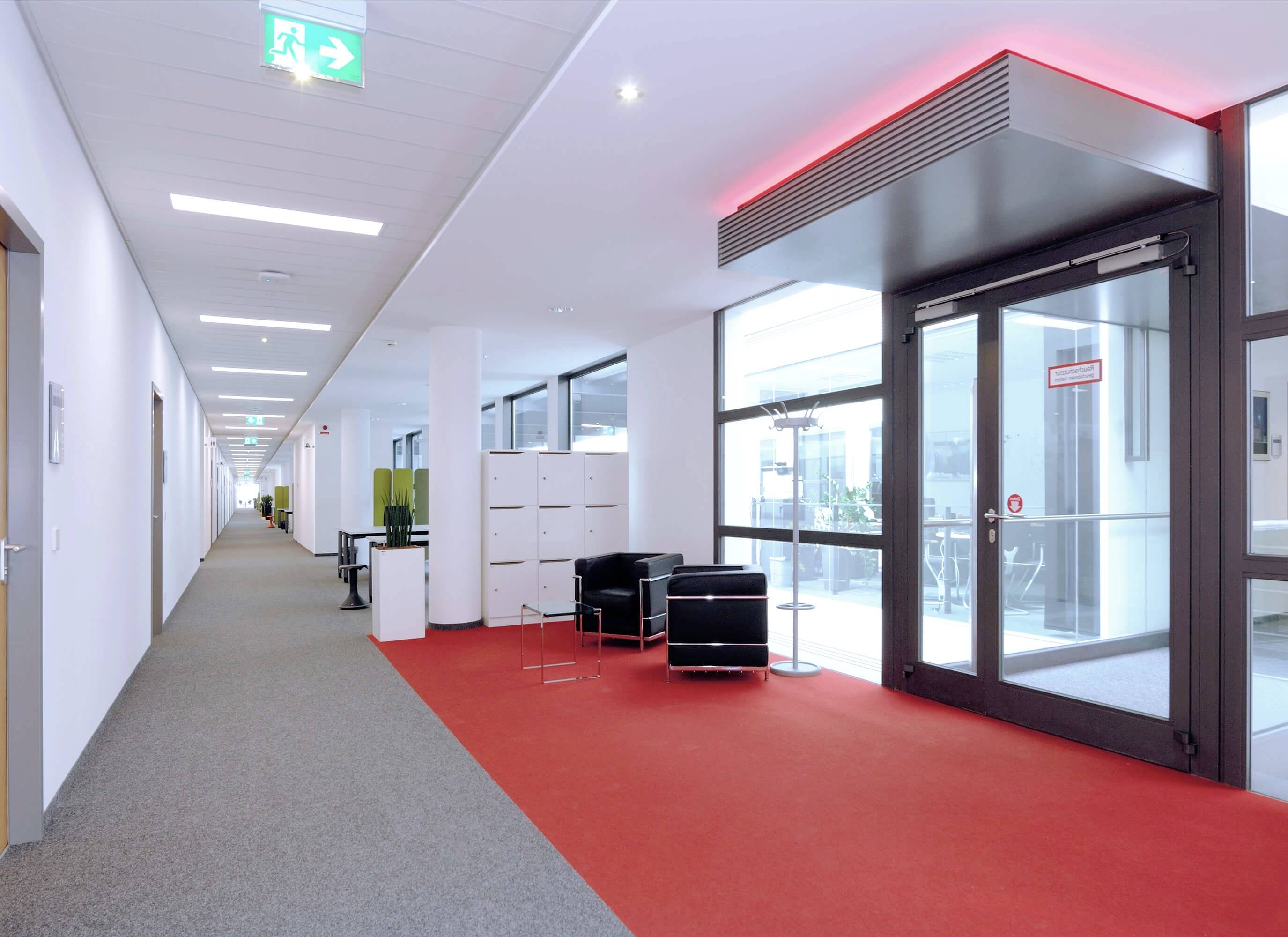 Bürokorridor mit rotem Teppich, Notausgangsschild an der Decke, Glasdoppeltür rechts, zwei schwarze Sessel und Tisch neben Schließfächern.