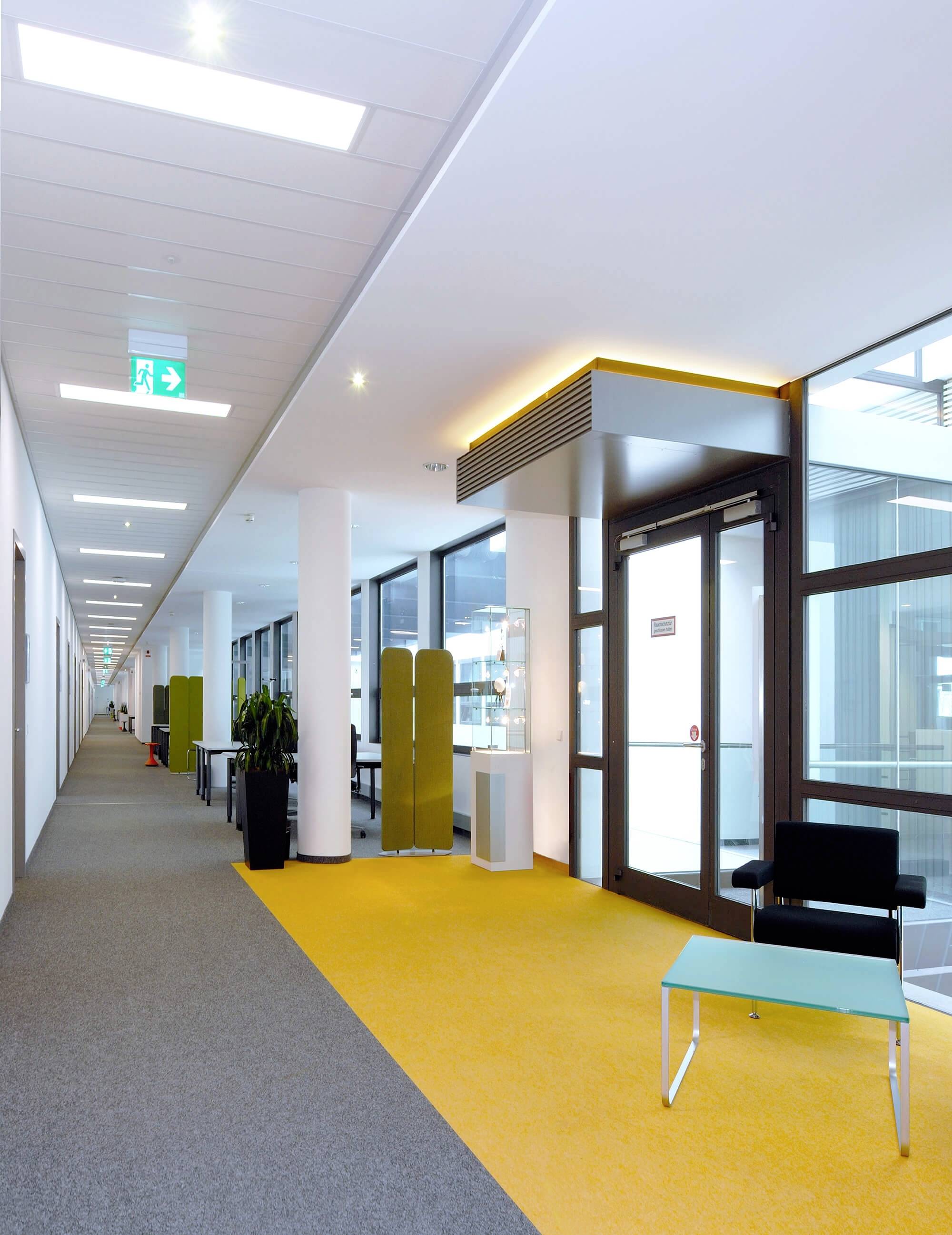 Moderner Büroflur mit gelbem Teppich, links Büropflanzen und Besprechungsnischen, rechts eine Glastür mit direktem Zugang zum Außenbereich.