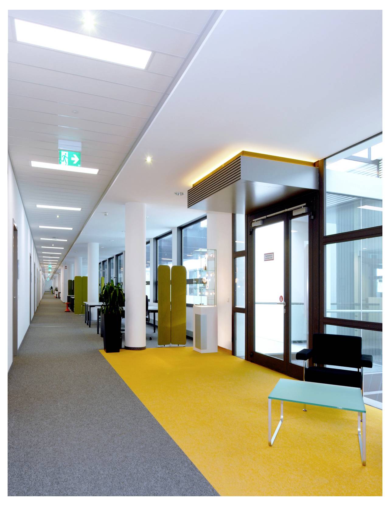 Moderner Bürokorridor mit grauem Teppichboden und gelb markiertem Bodenbereich, Räumen mit Glaswänden, einem schwarzen Stuhl und einem kleinen türkisfarbenen Tisch in der Nähe des Eingangs.