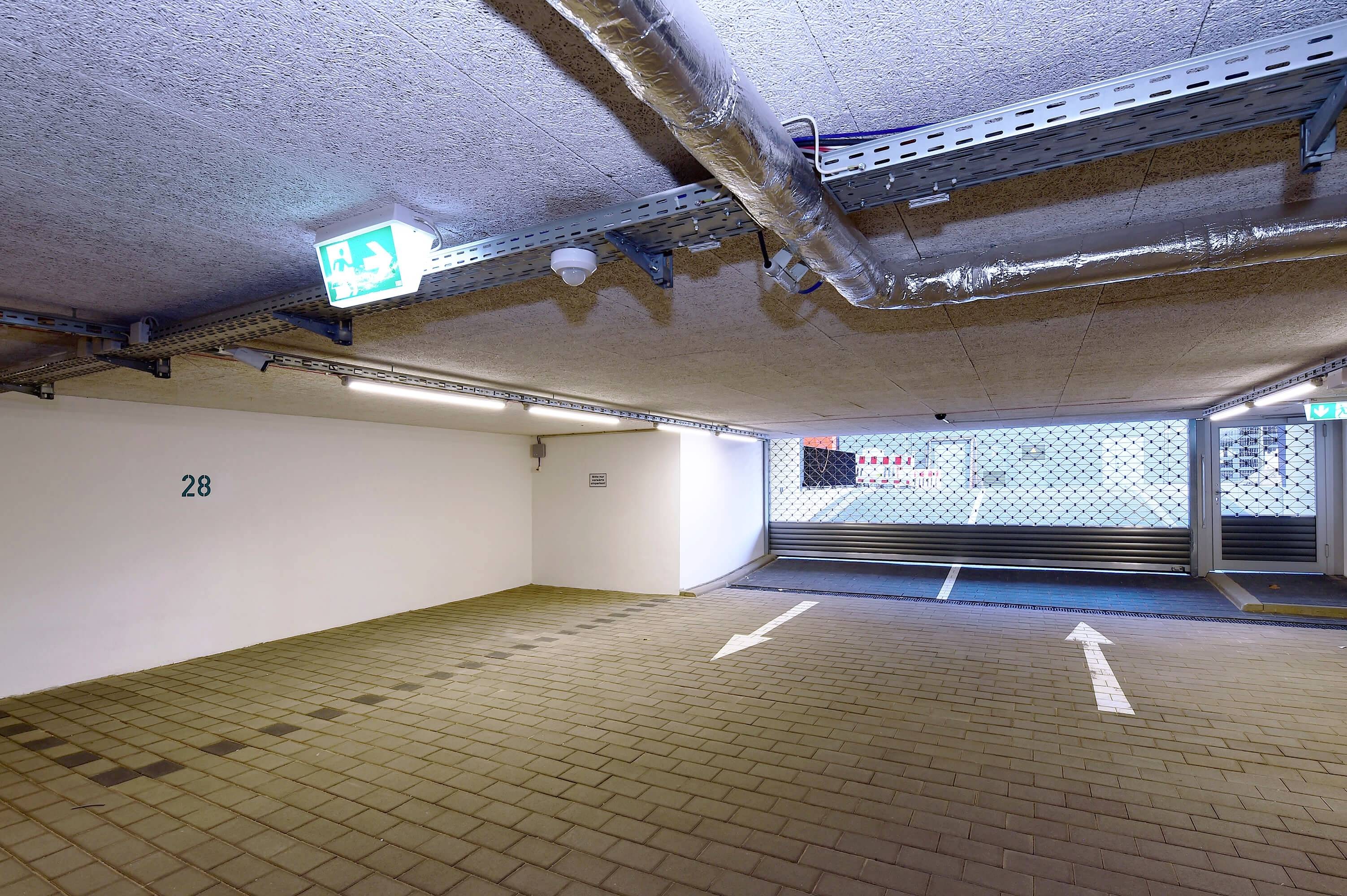 Ein leerer Parkplatz in einer Tiefgarage mit einem Notausgangsschild an der Decke und Richtungspfeilen auf dem Boden.