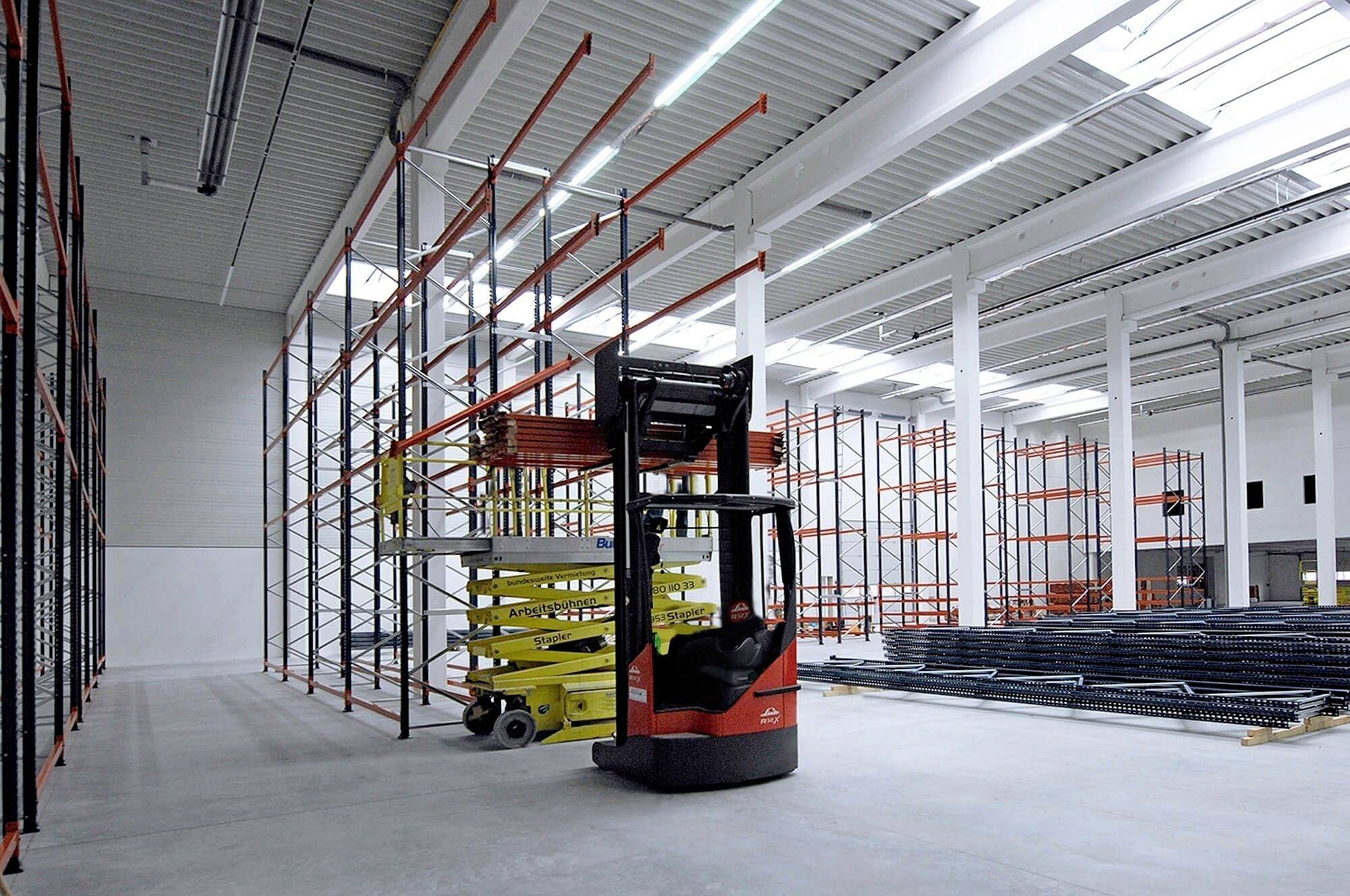 Gabelstapler hebt Metallgitter in einer leeren Lagerhalle, umgeben von leeren Regalen unter Tageslicht durch große Fenster.
