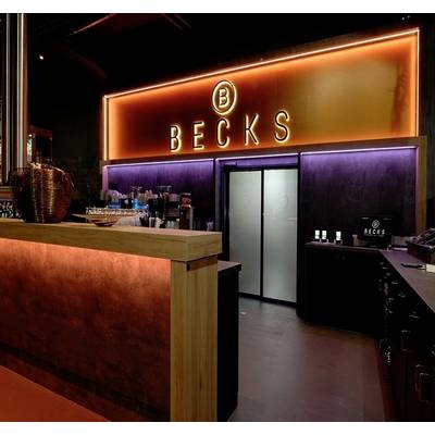 Bar mit hellem, orangefarbenem Neon-Schild 'BECKS' über dem Eingang. Dunkle Innenausstattung, Getränkebar links, moderne Atmosphäre.