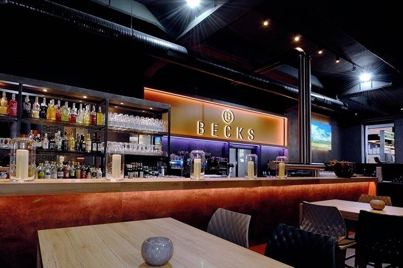 Eine moderne Bar mit beleuchtetem 'BECKS' Schild, gefüllten Regalen mit Flaschen und Gläsern. Holztheken und stilvolle Beleuchtung schaffen eine einladende Atmosphäre.