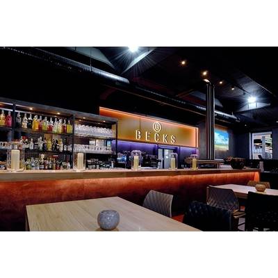 Eine moderne Bar mit beleuchtetem 'BECKS' Schild, gefüllten Regalen mit Flaschen und Gläsern. Holztheken und stilvolle Beleuchtung schaffen eine einladende Atmosphäre.
