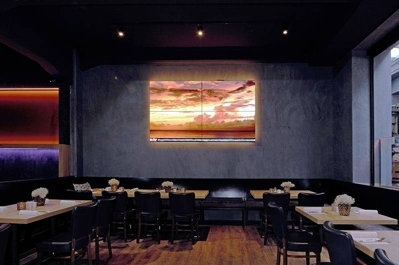 Restaurant mit dunkler Einrichtung, Holztischen und Stühlen. An der Wand ein beleuchtetes Wandbild mit Sonnenuntergang.
