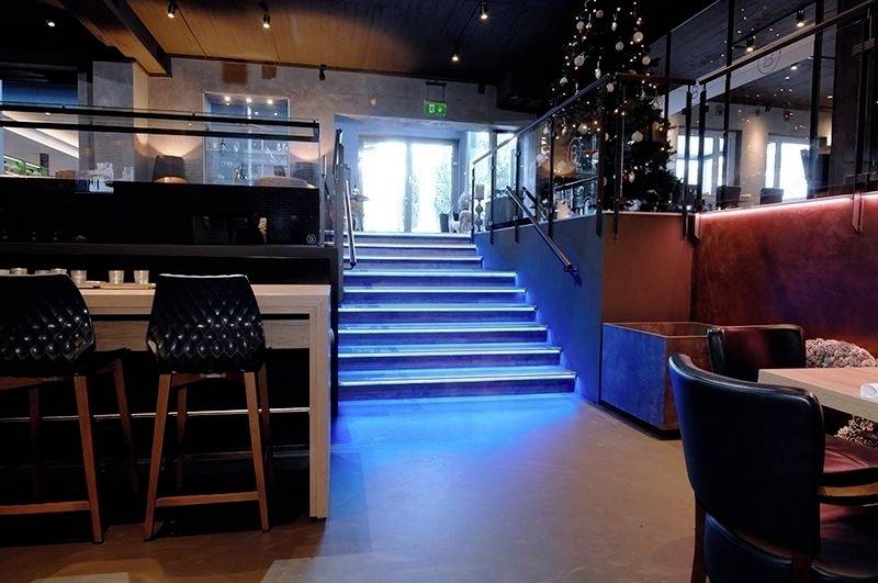 Eine modern eingerichtete Bar mit beleuchteter Treppe, die zu einer Glastür führt. Links sind Barstühle, rechts ein Weihnachtsbaum.