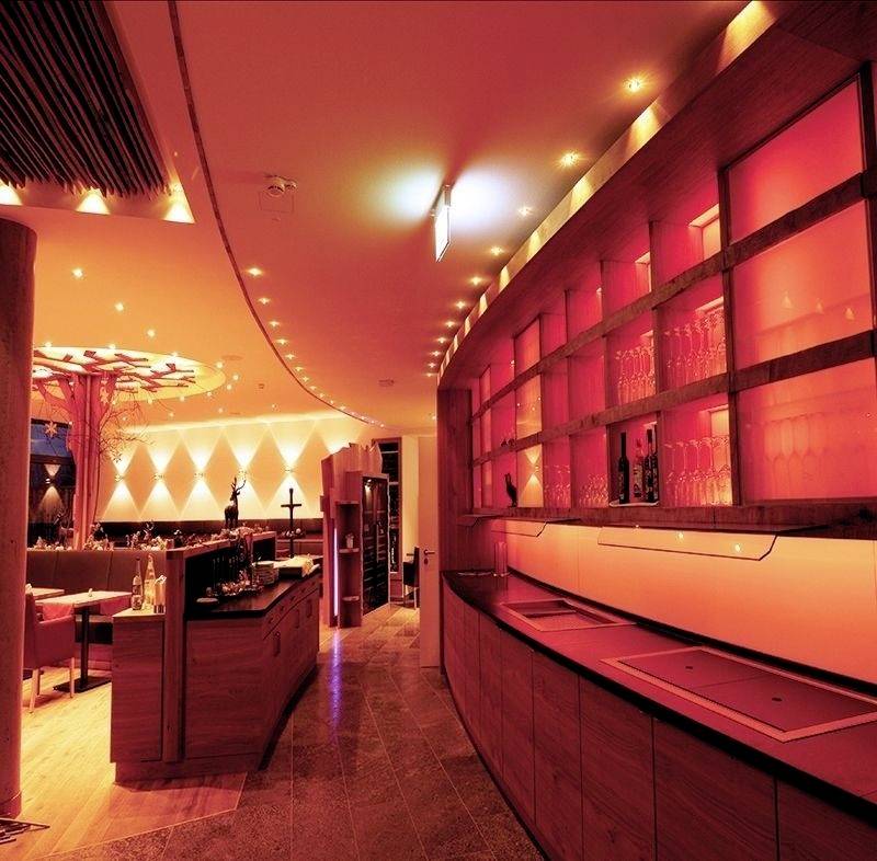 Eine moderne Bar mit rot-orange Beleuchtung und Holzmöbeln, einer langen Theke links und Regalen mit Gläsern an der rechten Wand.