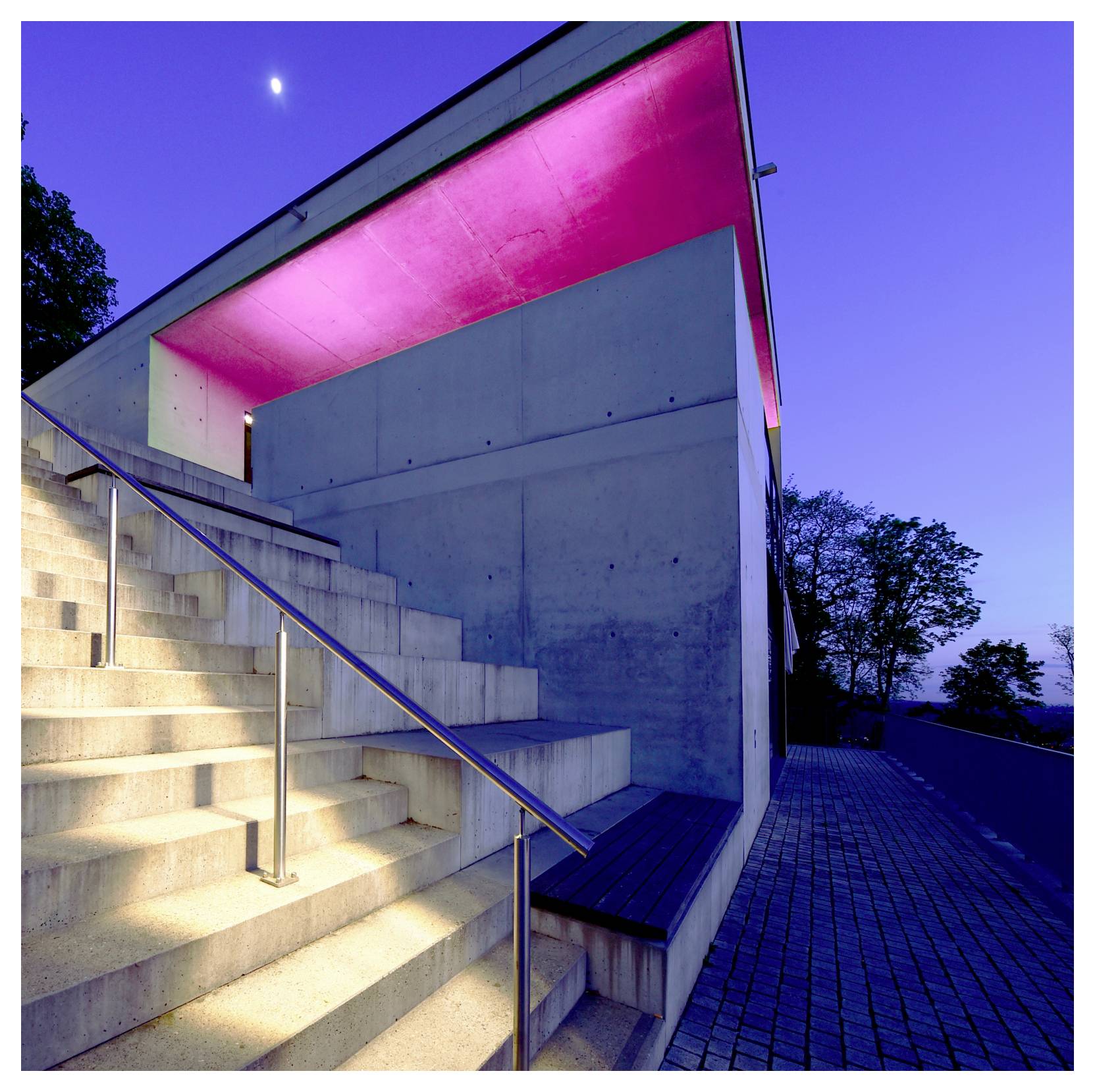 Modernes Betongebäude mit beleuchtetem Treppenhaus und leuchtend pinkfarbenem Dach unter dem Abendhimmel.