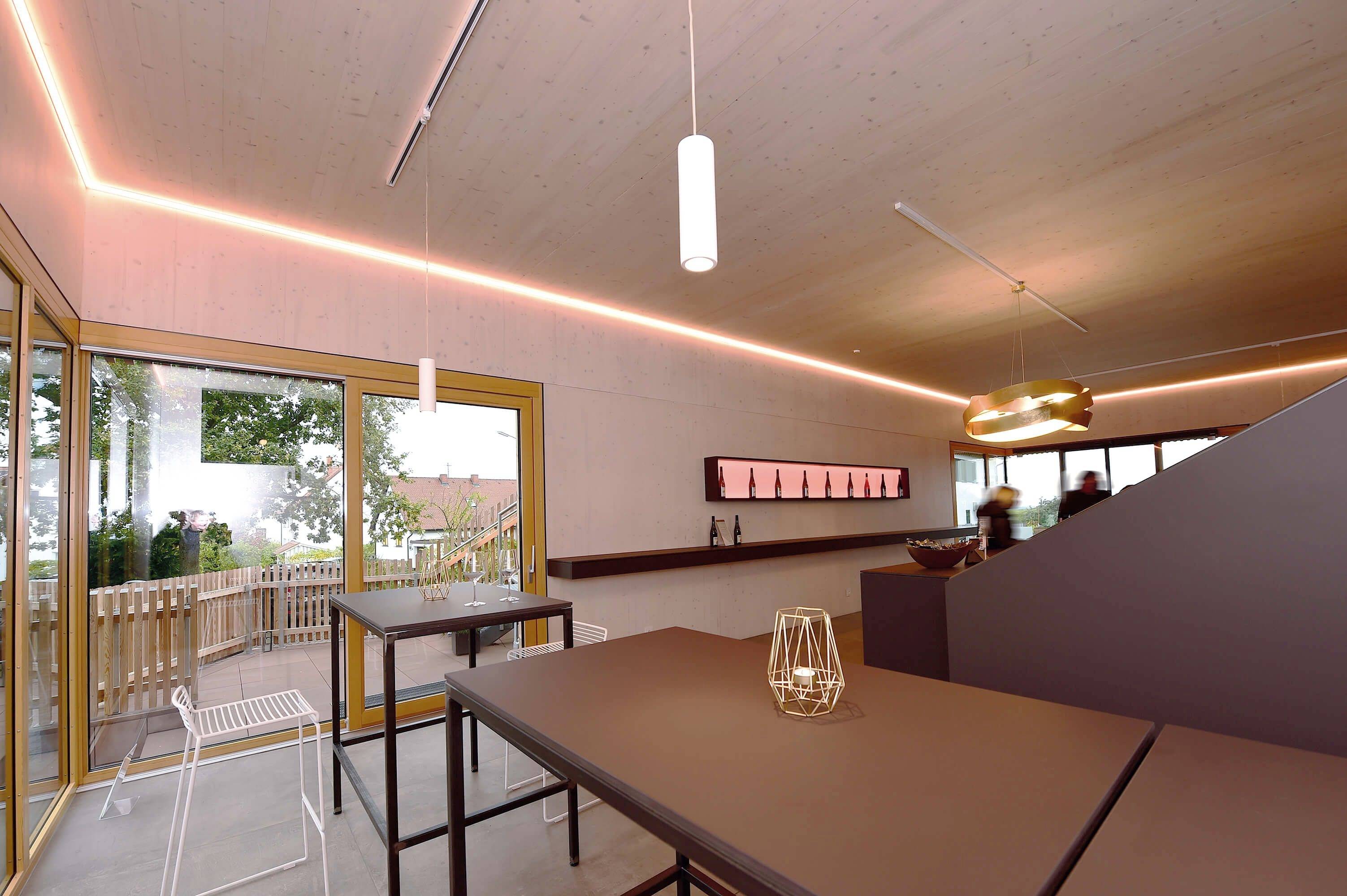 Moderner Innenraum eines Café mit Holzverkleidung, stilvollen Tischen, Hockern und großen Fenstern. Außenbereich mit Holzterrasse erkennbar.