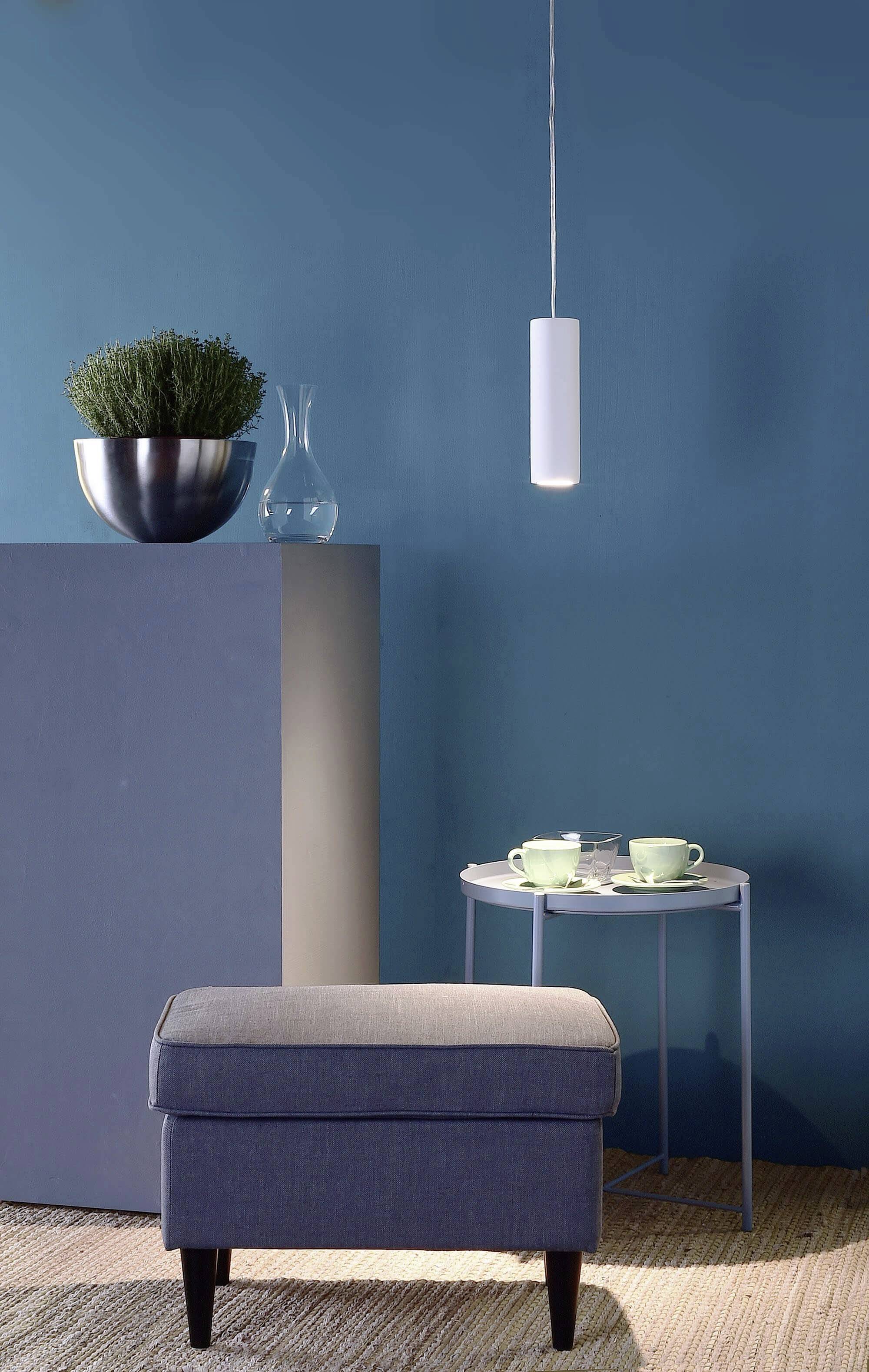 Ein Zimmer mit blauem Hintergrund, moderner Hängelampe, zwei Tassen auf kleinem Tisch, grauer Polsterhocker und Pflanze in Metalltopf.