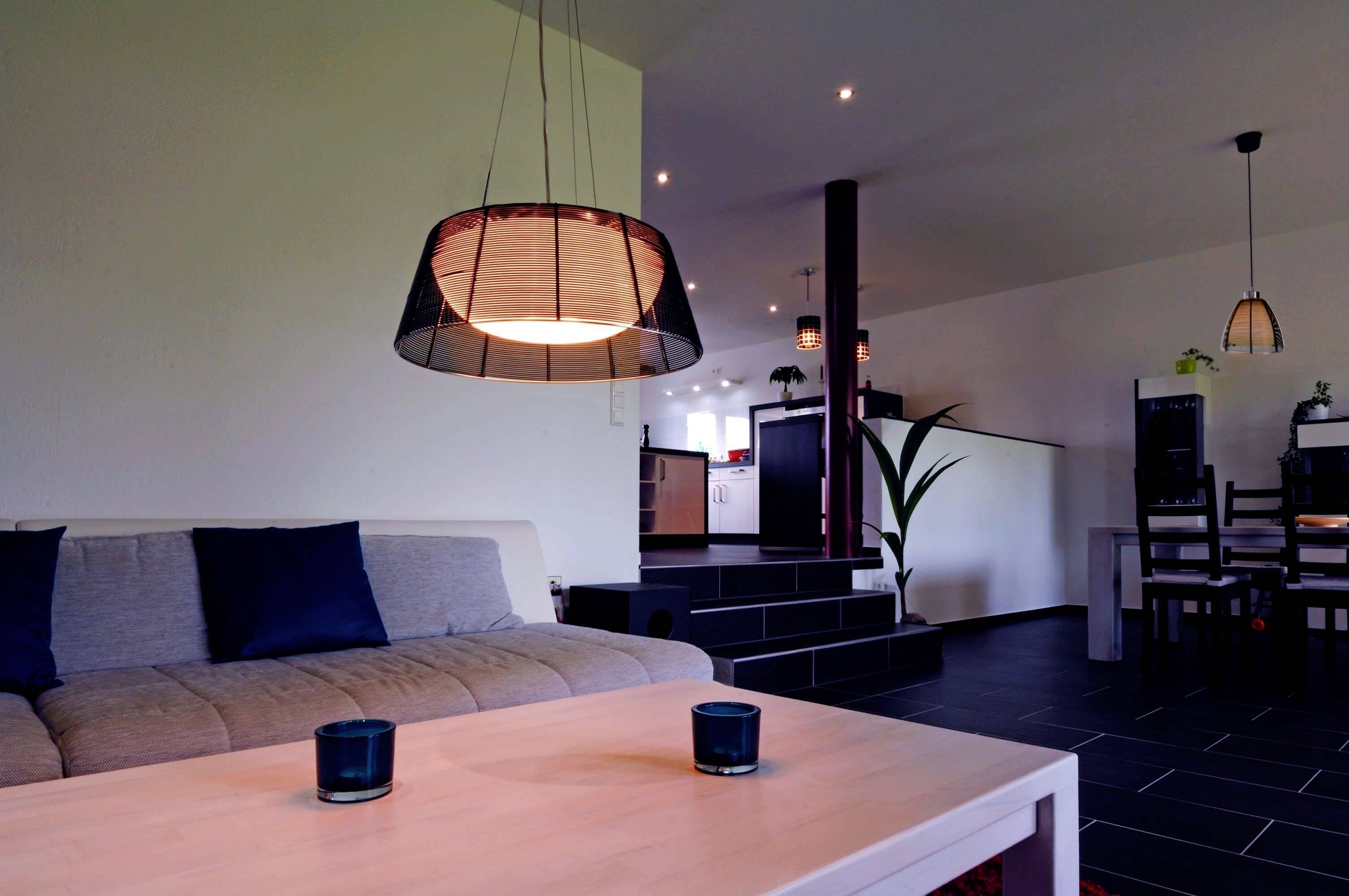 Modernes Wohnzimmer mit grauem Sofa und blauen Kissen, Holz-Tisch und hängenden Lampen. Offene Küche im Hintergrund.