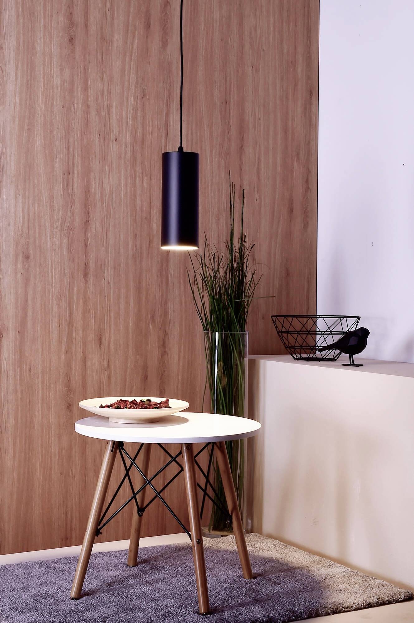 Ein runder, weißer Tisch mit Holzwangen steht auf einem Teppich. Darüber hängt eine schwarze Pendelleuchte. Dekorative Pflanzen im Hintergrund.