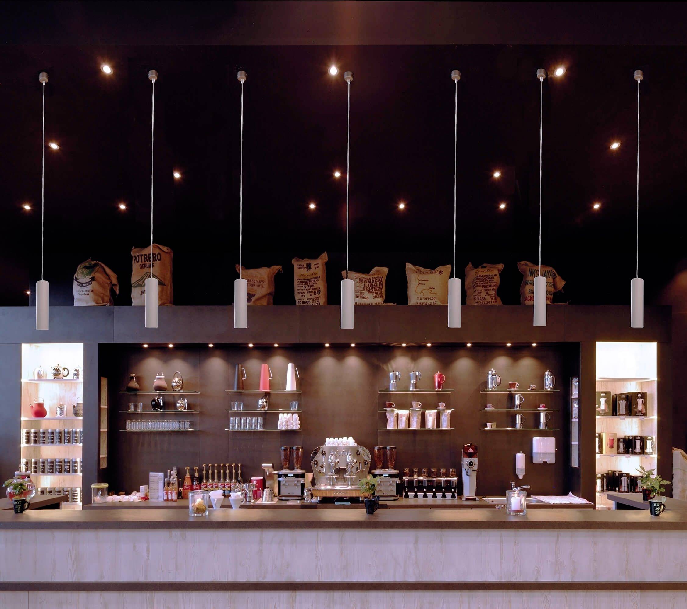Eine moderne Café-Theke mit hängenden Lichtern, Kaffeemaschine, Tassen und Kaffeesäcken im Hintergrund. Gemütliche Atmosphäre.