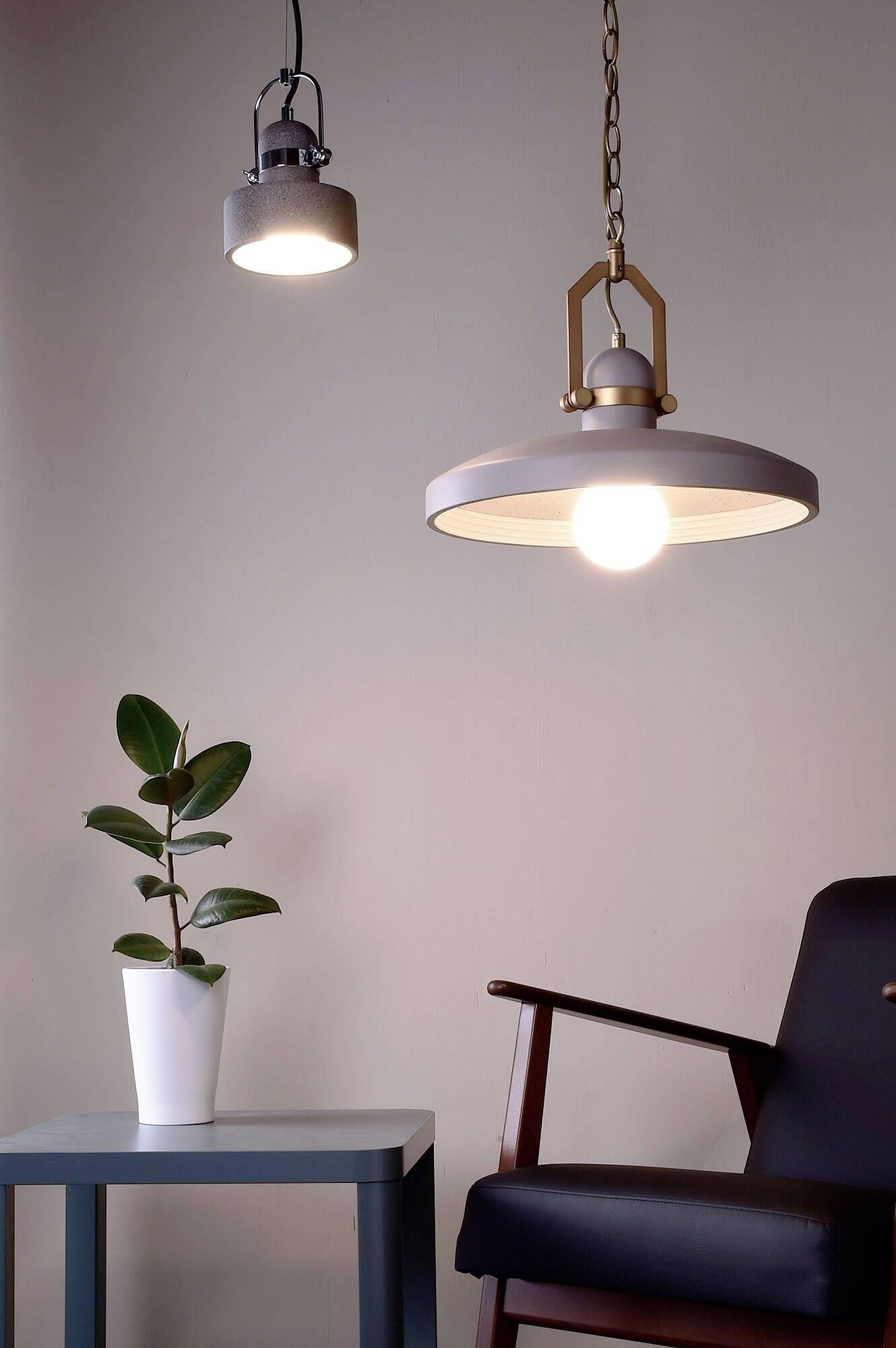 Eine moderne Zimmerlampe hängt über einem Tisch mit einer Pflanze in einem weißen Topf. Daneben steht ein hölzerner Stuhl.