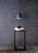 Ein minimalistischer Raum mit einer hängenden Lampe über einer modernen Skulptur auf einem schmalen Tisch vor einer grauen Wand.