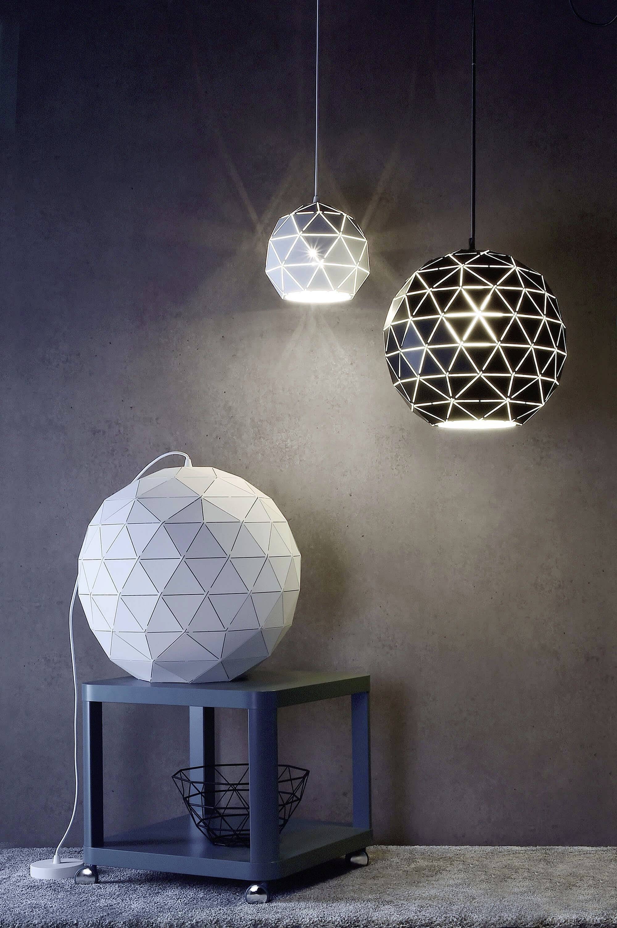Zwei hängende geometrische Lampen, eine weiß und eine schwarz, erhellen einen Raum. Eine ähnliche Tischlampe steht auf einem kleinen Tisch.