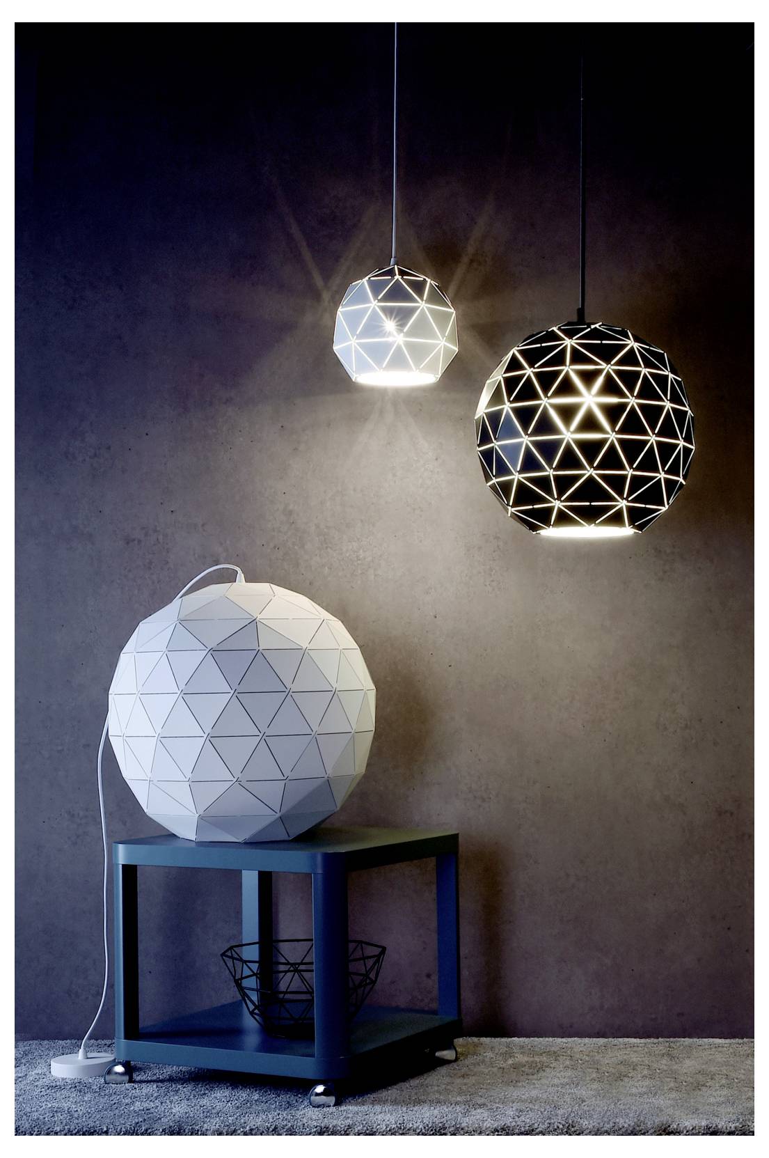 Drei geometrische Pendelleuchten hängen über einer weißen, facettierten Lampe auf einem blauen Tisch vor einer dunklen Wand.