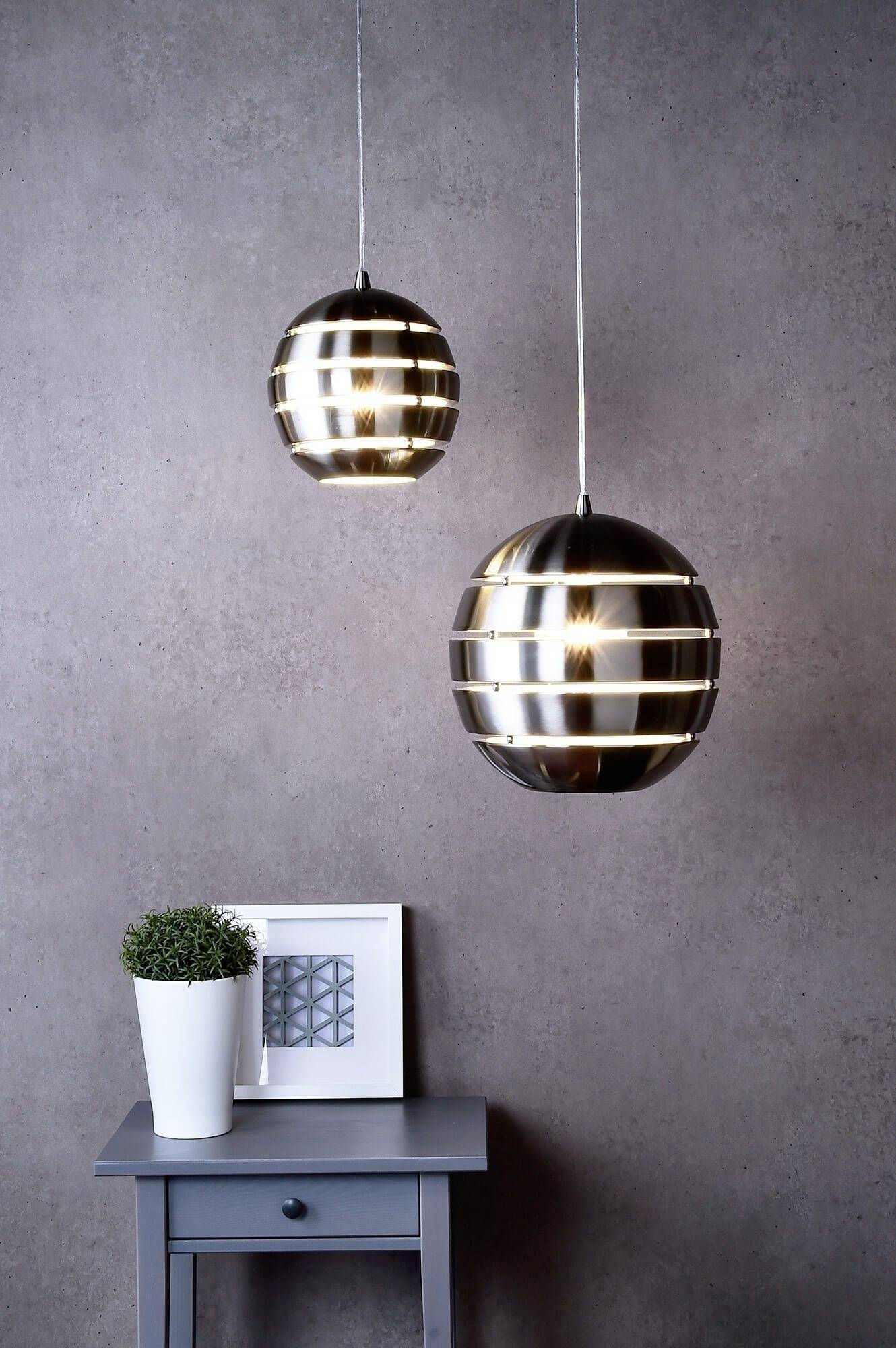 Zwei hängende, metallisch glänzende Lampen über einem kleinen Tisch mit Pflanze und gerahmtem Bild. Rustikale Wand im Hintergrund.