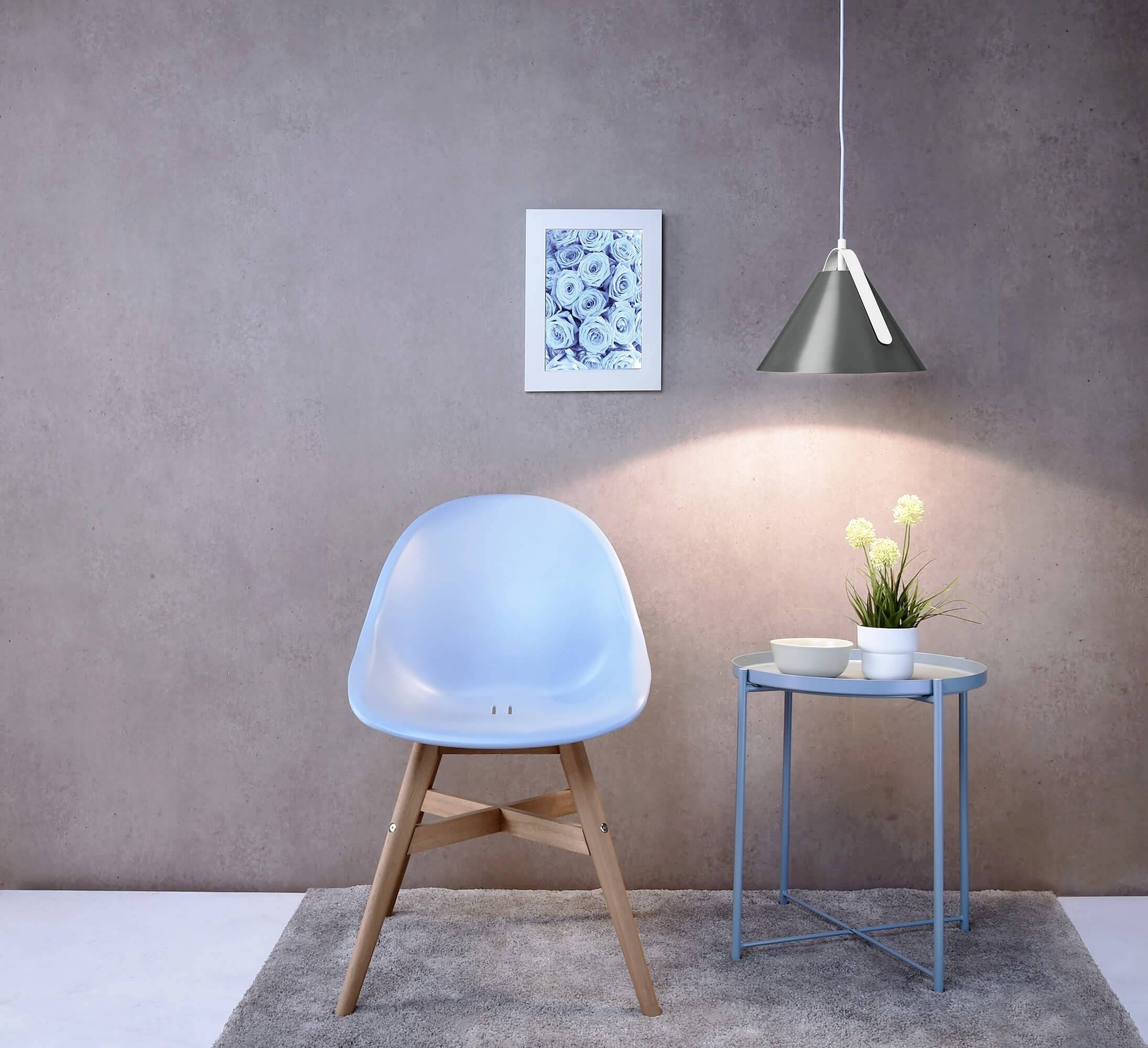 Ein blauer Stuhl steht auf einem grauen Teppich vor einer Wand. Rechts daneben ein kleiner Tisch mit einer Pflanze und zwei Tassen, beleuchtet von einer Hängelampe.