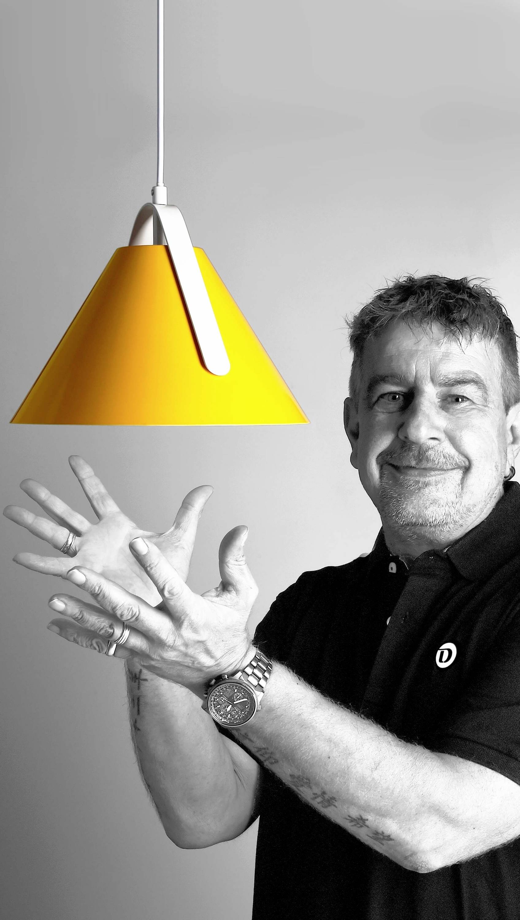 Ein Mann mit kurzem Haar und Tätowierungen hält eine leuchtend gelbe Lampe. Er trägt ein schwarzes Polohemd und lächelt leicht.