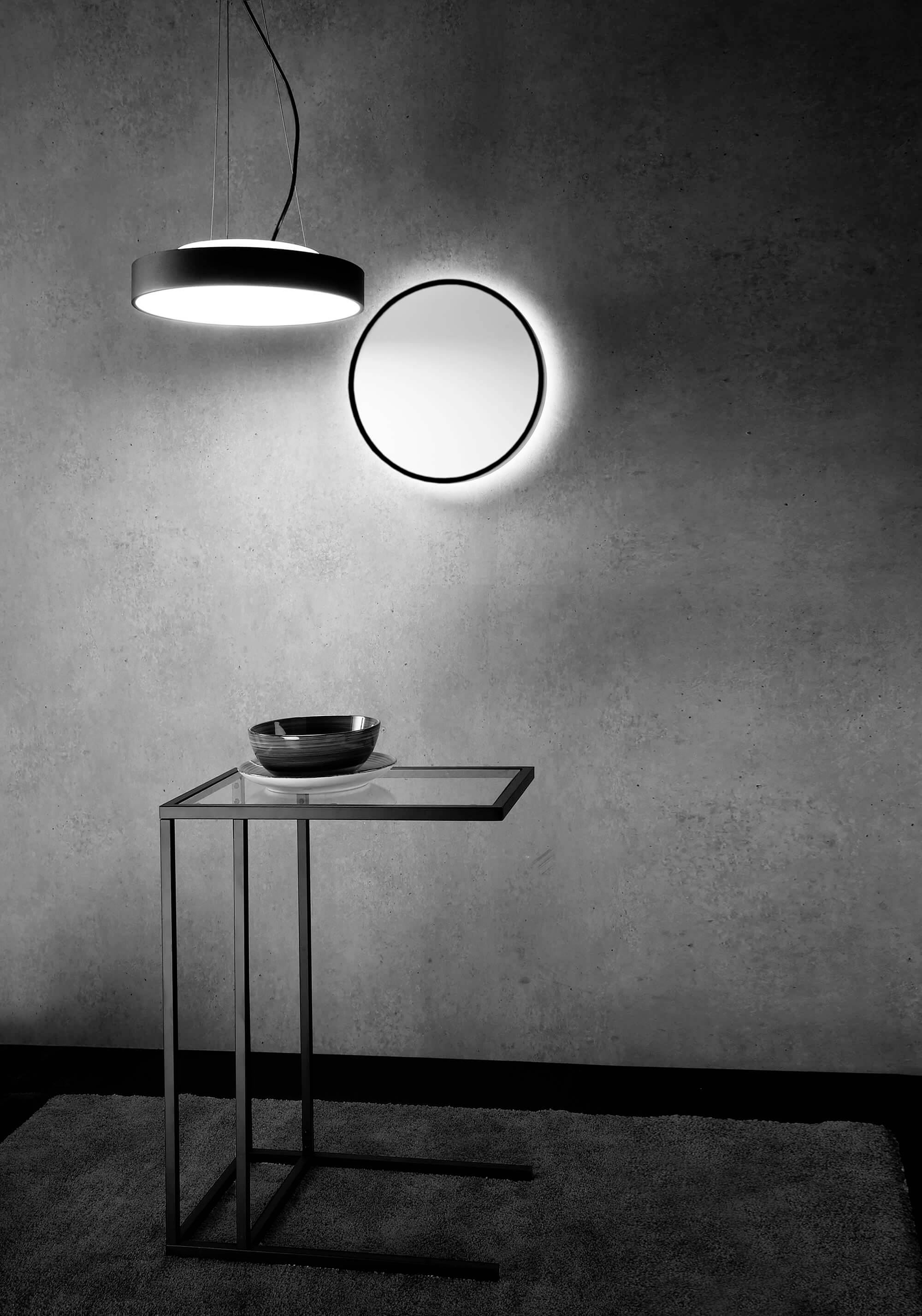 Ein minimalistisches Interieur mit einem runden Spiegel an der Wand, einer hängenden Lampe darüber, einem Glastisch und einer Schale darunter.