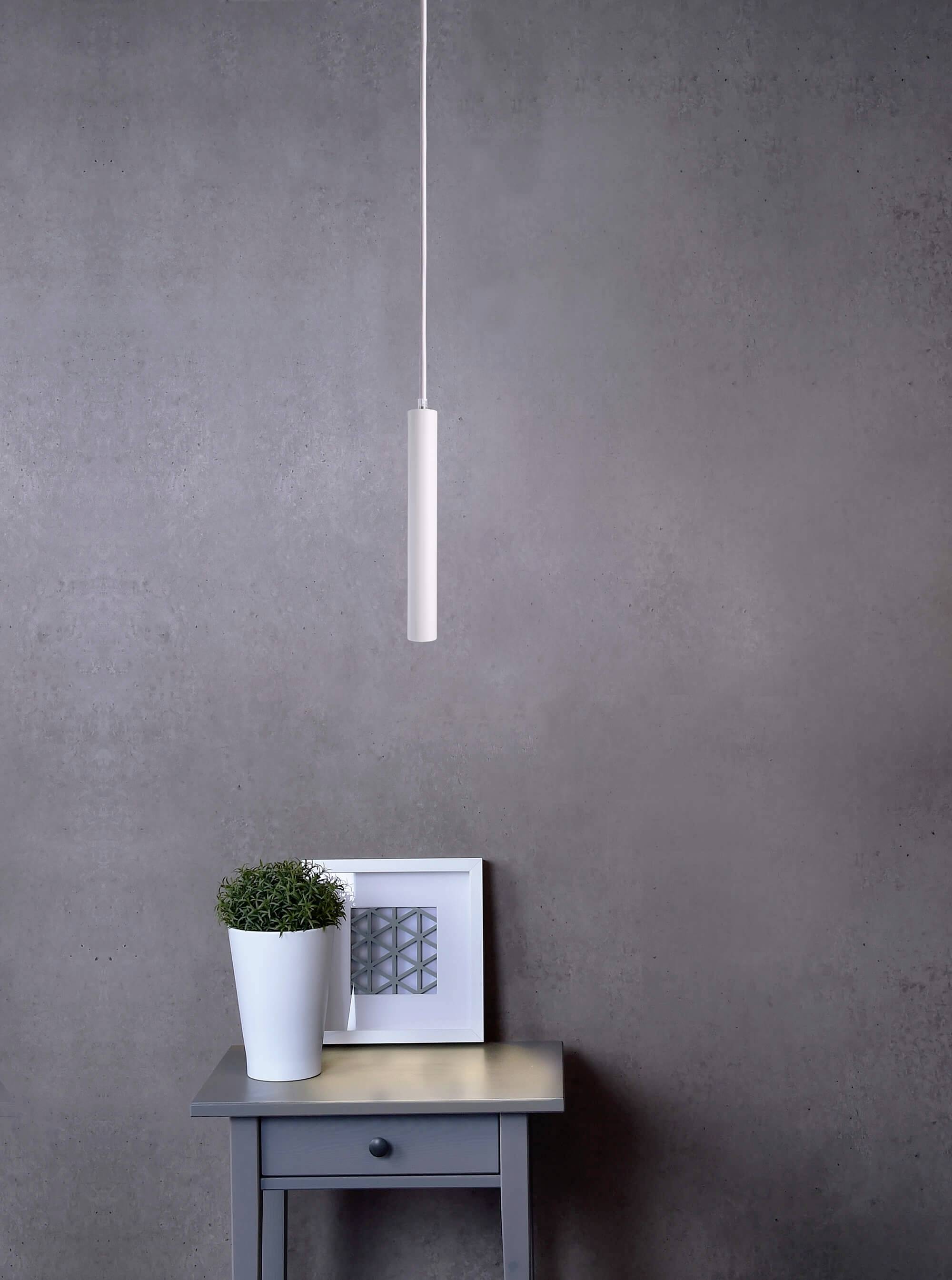 Eine graue Wand mit einer hängenden, schlanken Lampe. Darunter steht ein kleiner Tisch mit einem weißen Blumentopf und einem gerahmten Bild.