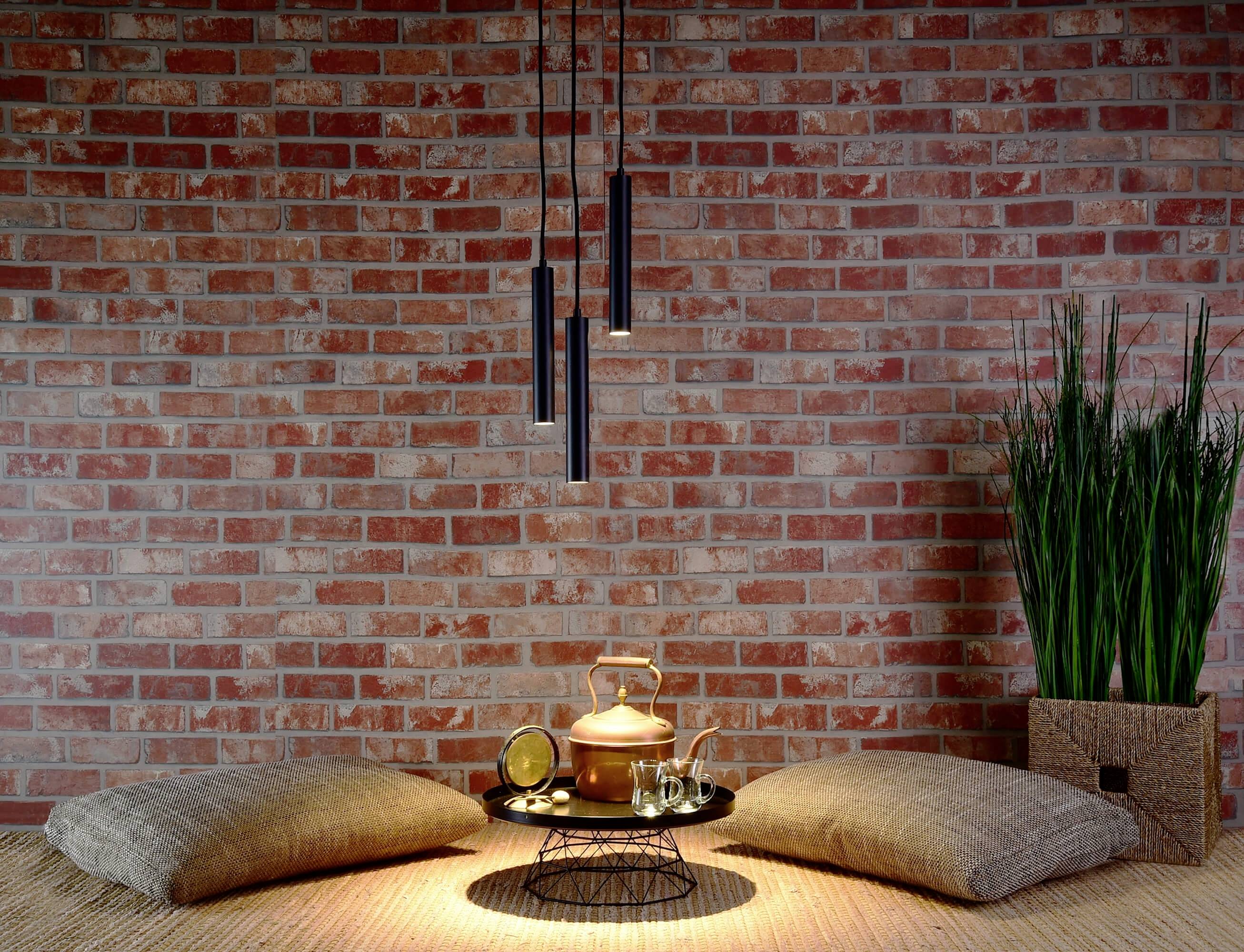 Ein Ziegelzimmer mit zwei Kissen am Boden, einem Teetablett mit Kessel und Tassen im Vordergrund, schlichte Lampe und Pflanzen rechts.